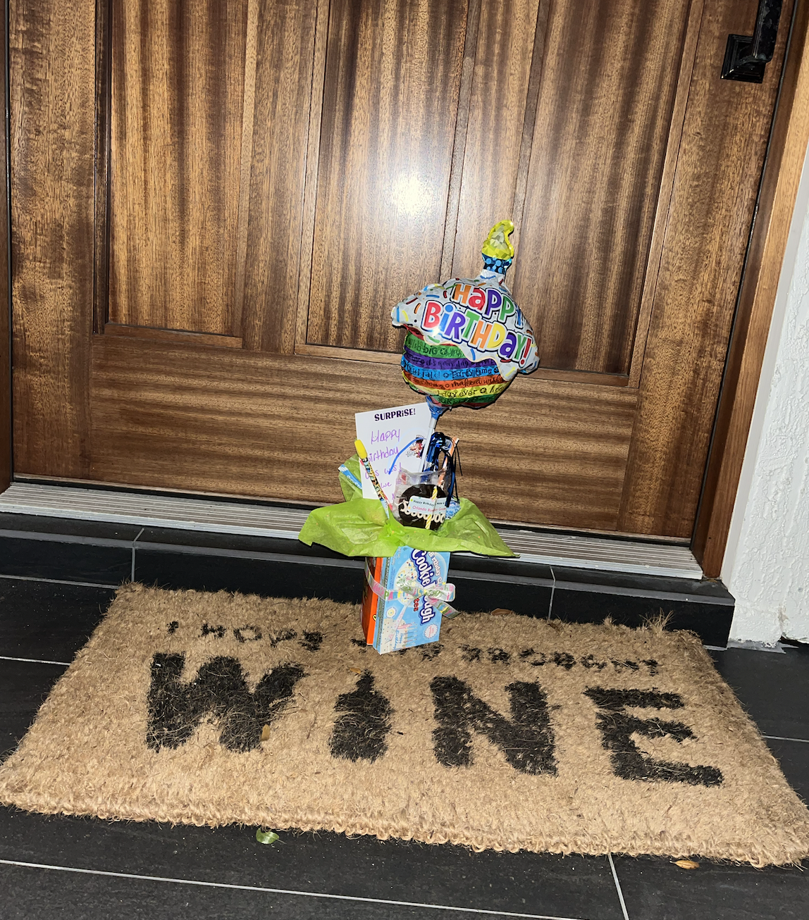 Birthday surprise setup with a colorful balloon bouquet, a Hello Kitty box, and a handwritten birthday card placed on a green napkin on a floor mat in front of a wooden door.