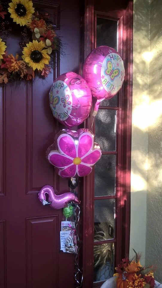 A cluster of balloons hanging on a red door, including two round 'Happy Birthday' balloons, a pink flower-shaped balloon, and a pink flamingo balloon, with a wreath of sunflowers and fall leaves on the left and sunlight casting shadows on the wall.