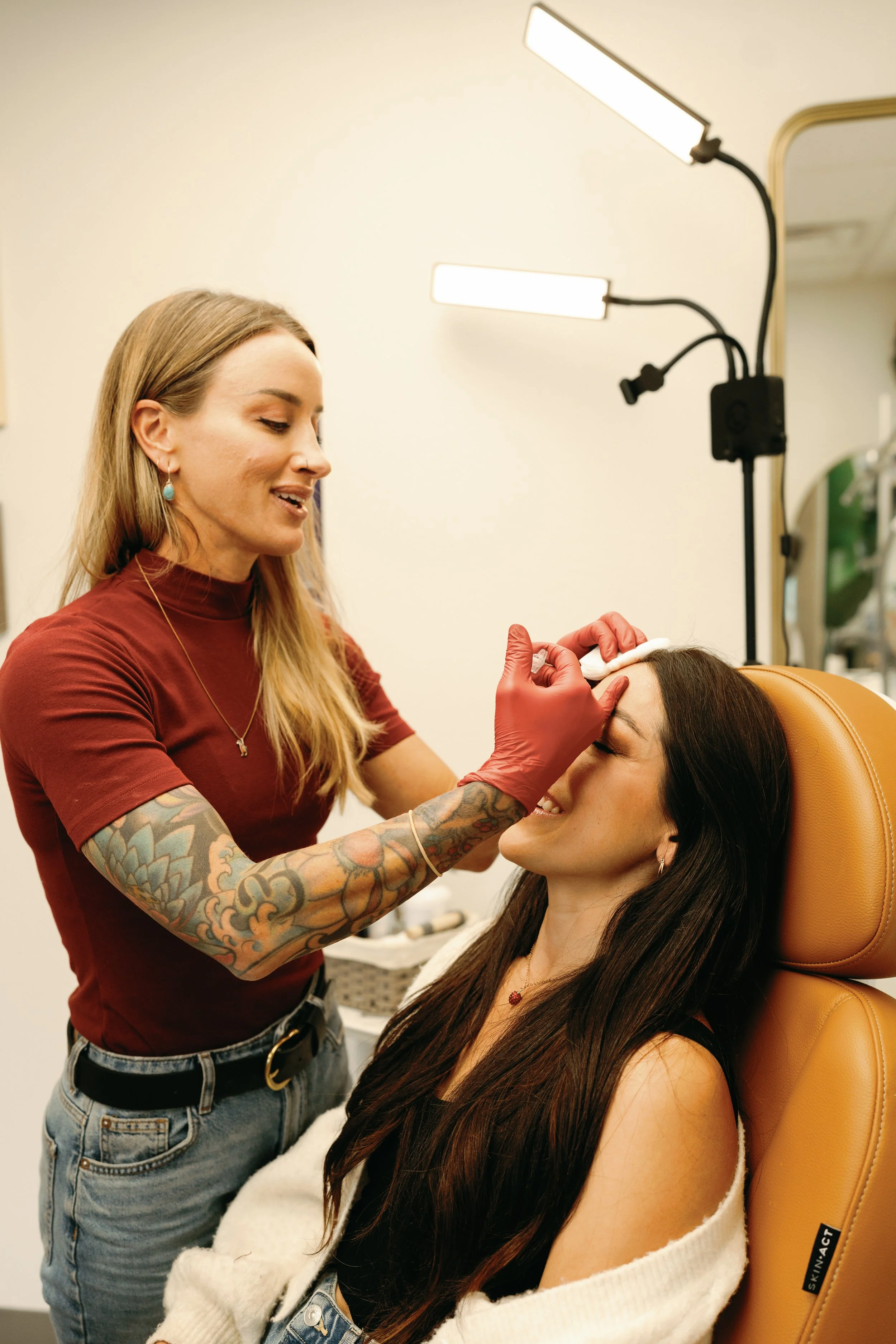 A woman with tattoos on her right arm providing a cosmetic or skincare treatment to another woman sitting in a chair, in a professional setting with lighting equipment surrounding them.