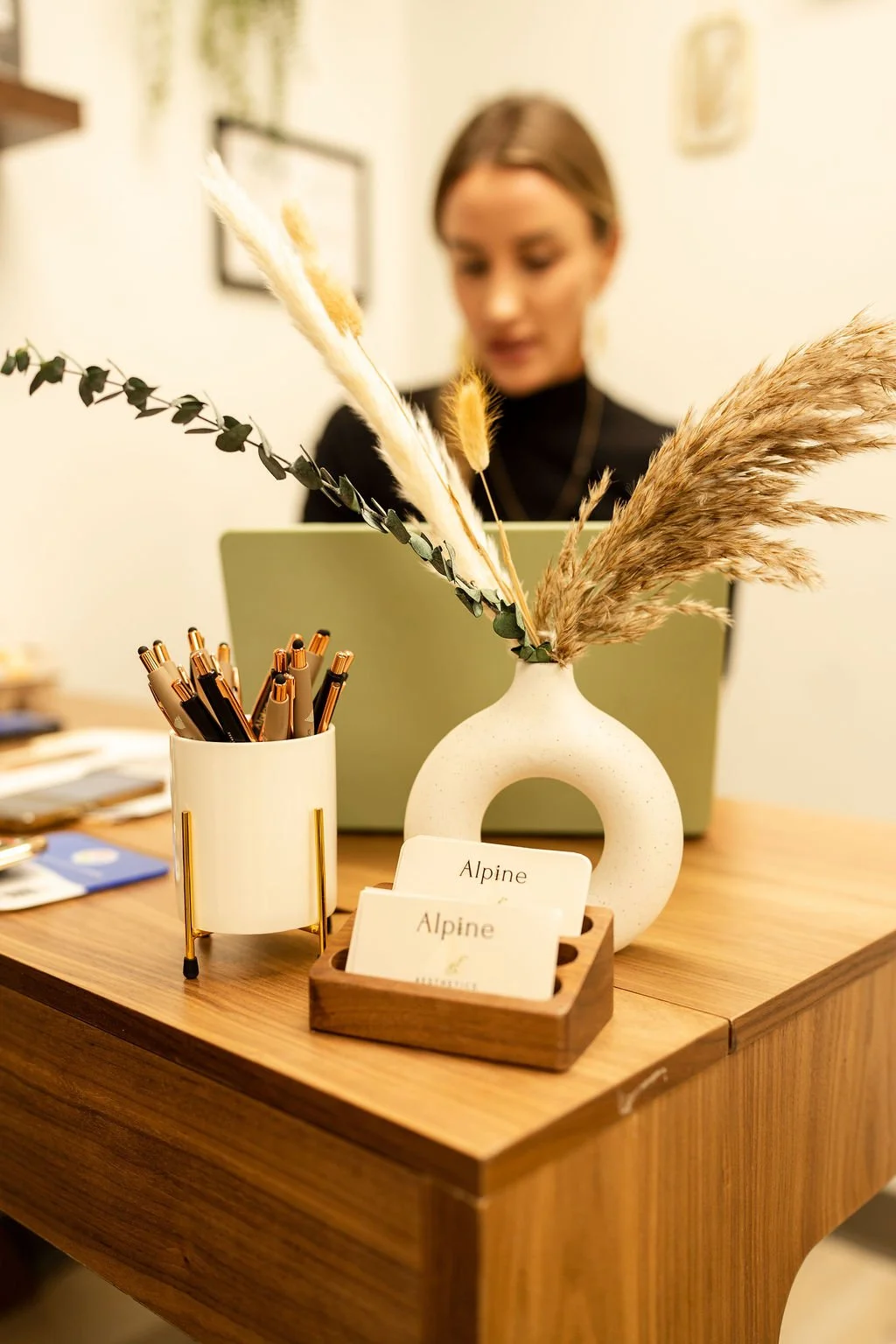 Close-up of a wooden desk with a beige vase holding dried pampas grass and eucalyptus, a white pen holder with gold pens, and a wooden cardholder with business cards labeled "Alpine." In the background, a woman with brown hair and a black top is working on a pale green laptop.