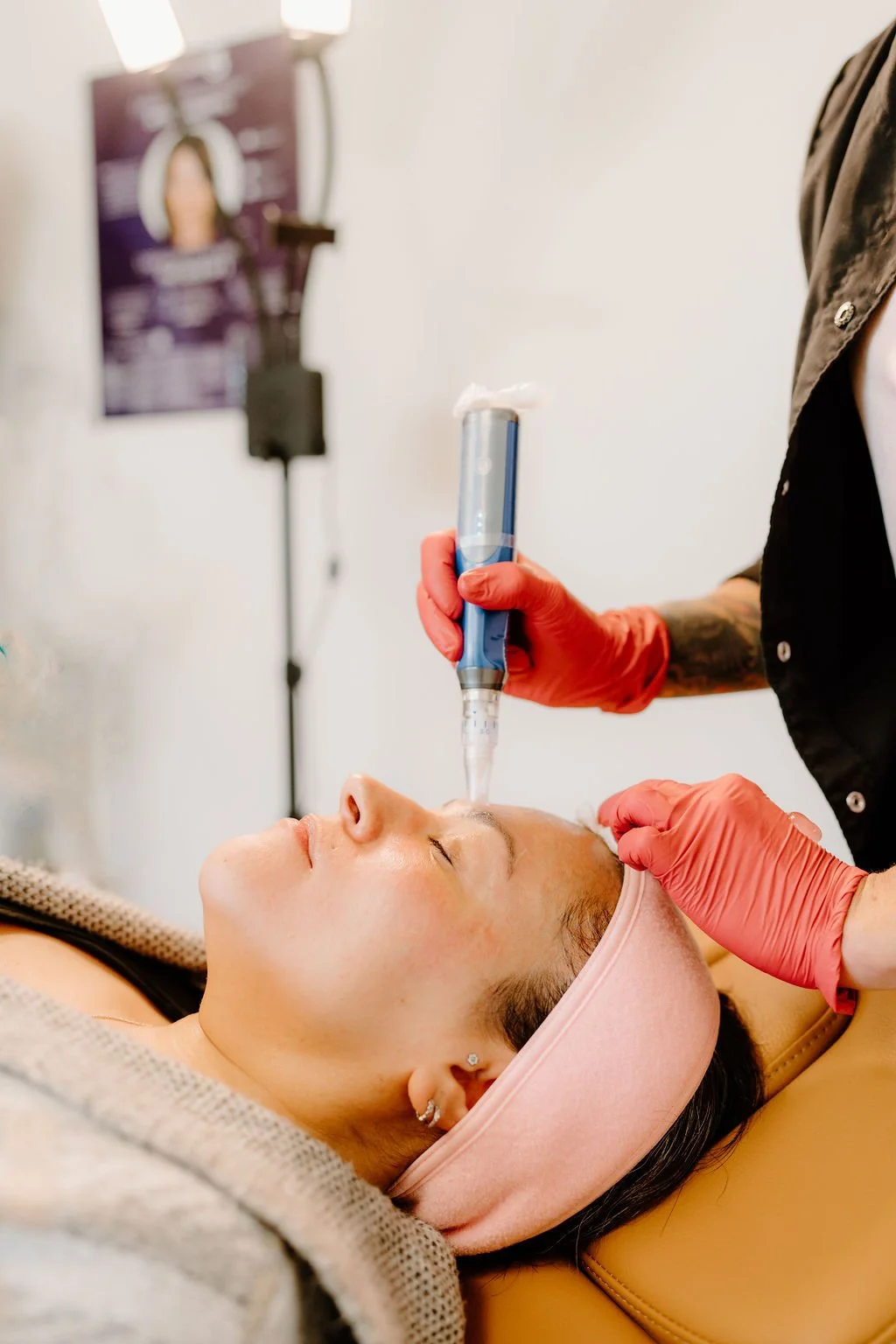 A woman lying down with her eyes closed while a skincare professional uses a handheld device on her face during a treatment in a clinic.