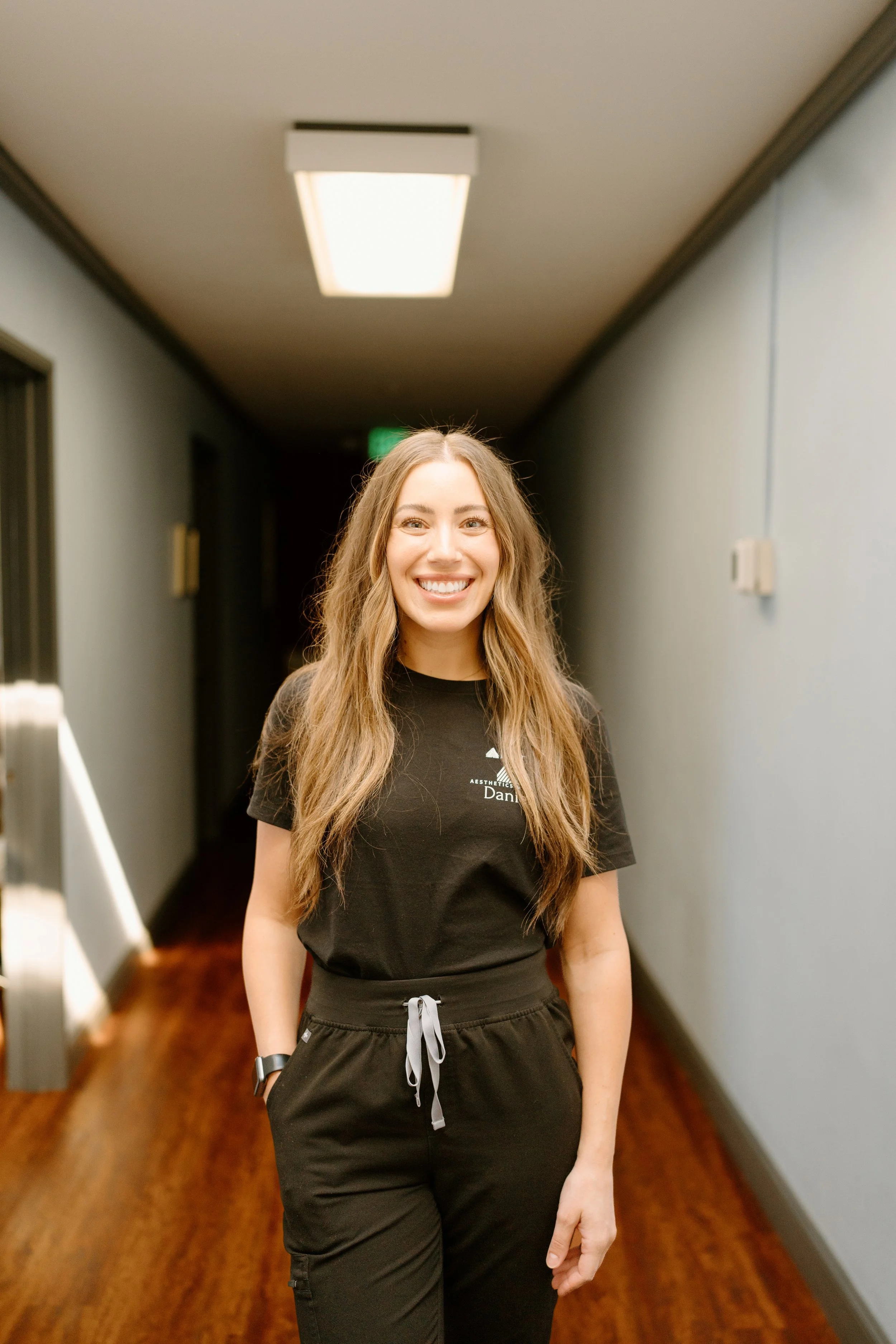 A young woman with long wavy hair smiling, standing in a hallway with gray walls and wooden flooring, illuminated ceiling light