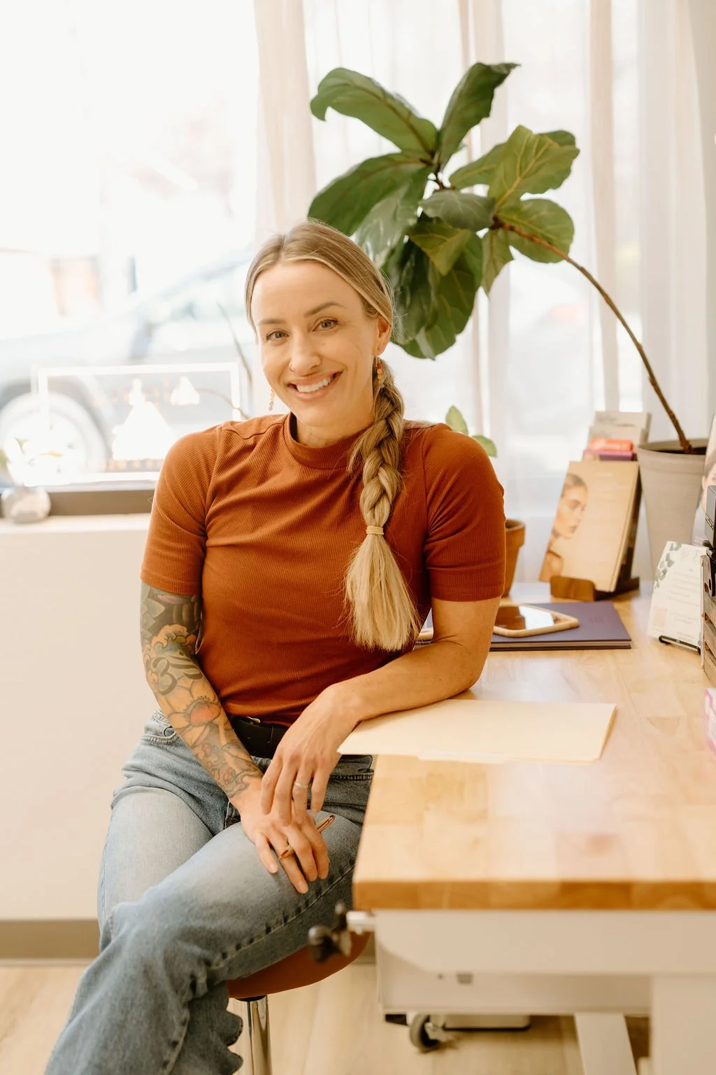 Smiling woman with long braided hair and tattoos on her arm sitting at a wooden desk in a brightly lit room with a large green plant and various papers and magazines.