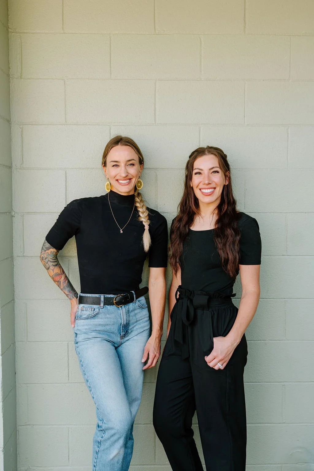 Two women in black tops and pants smiling and standing against a light green brick wall.