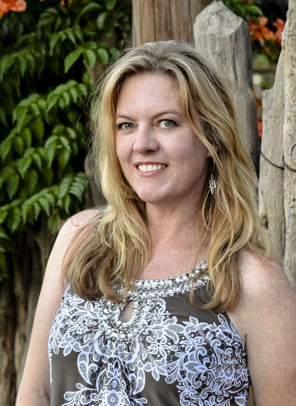 A woman with long blonde hair and blue eyes, smiling, wearing a sleeveless dress with white and silver embellishments, standing outdoors near a wooden fence and green foliage.