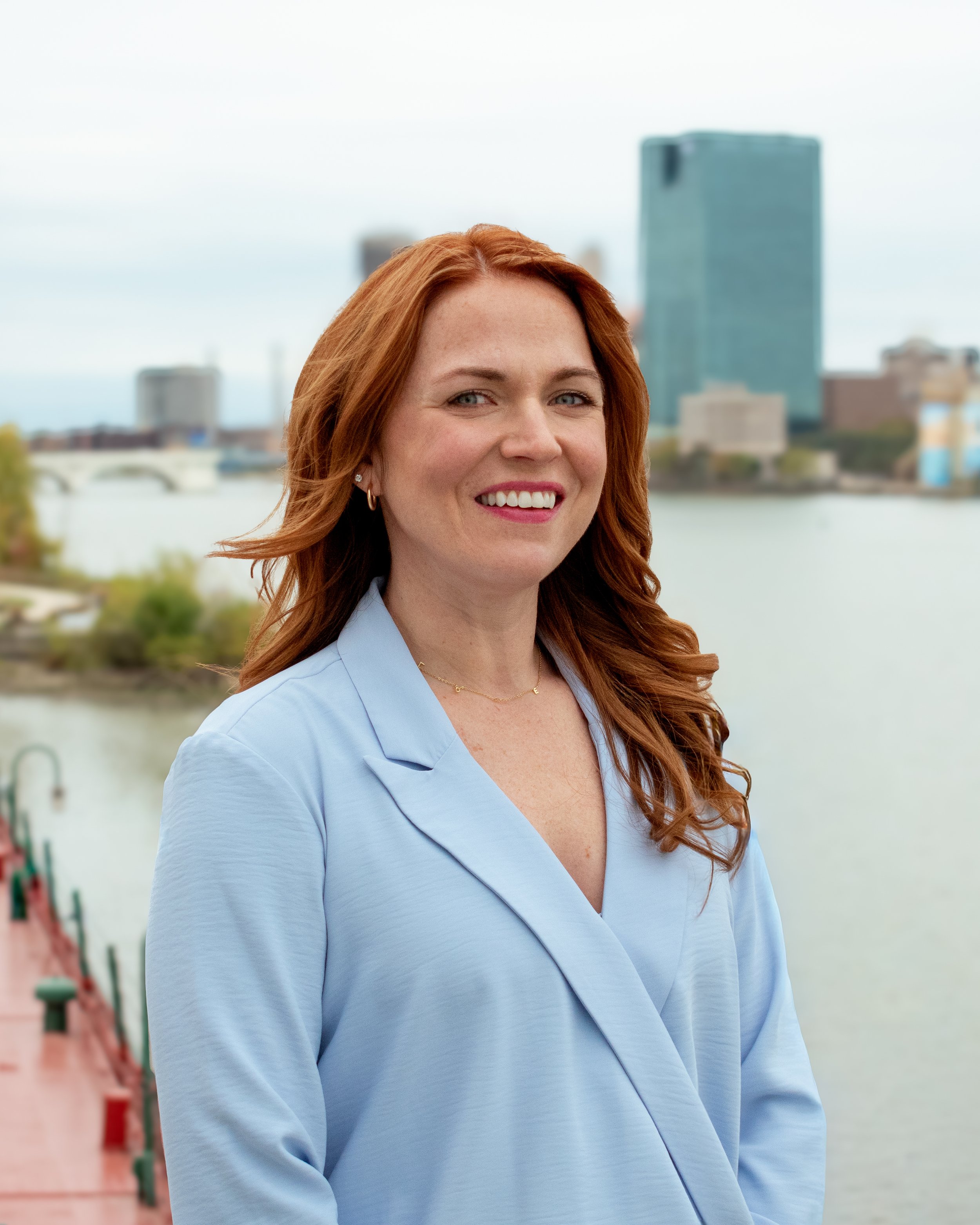 A woman with red hair, wearing a light blue shirt, smiling outdoors in an urban setting with buildings and a river in the background.