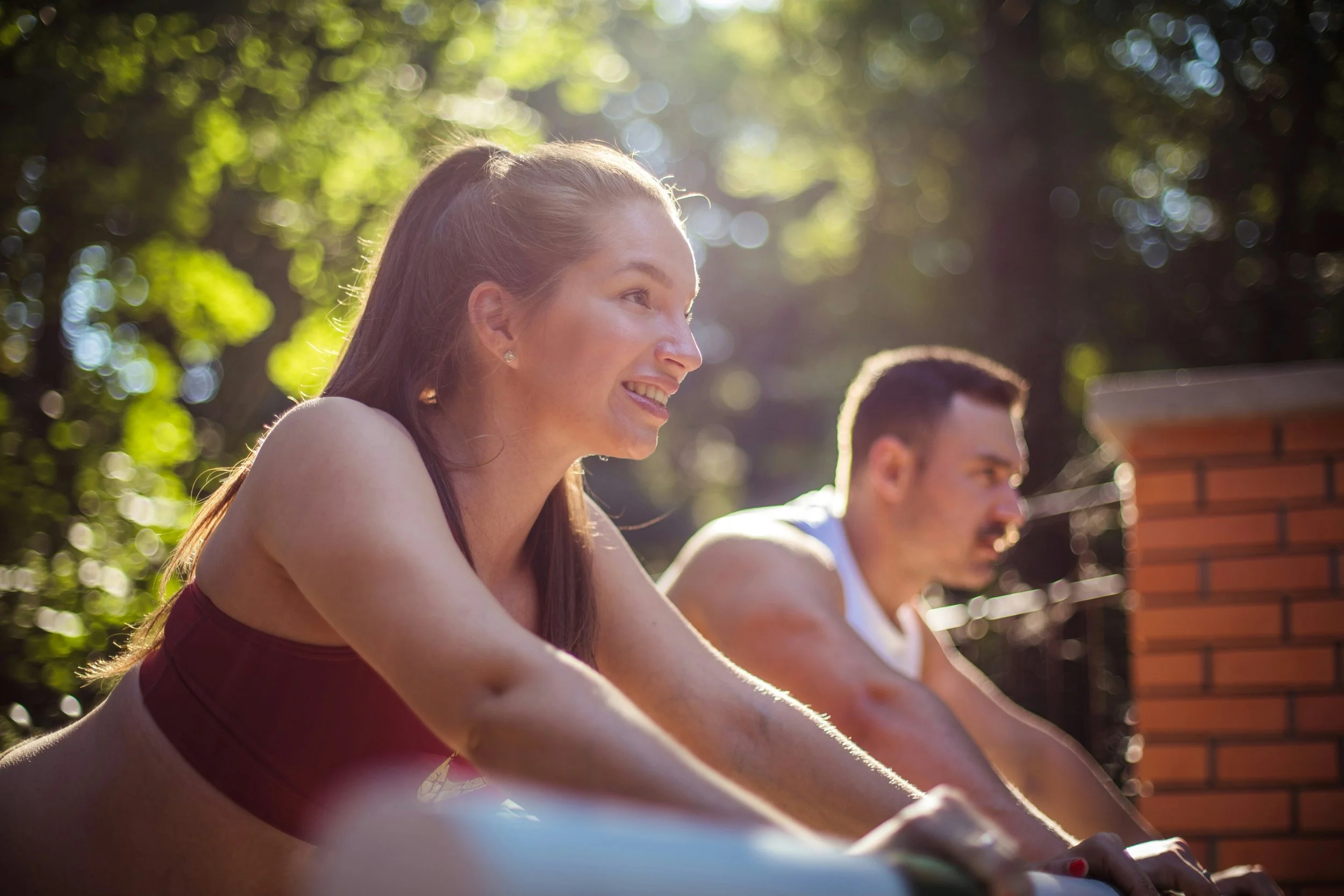 Three people sitting outdoors during daytime with sunlight filtering through trees in the background.