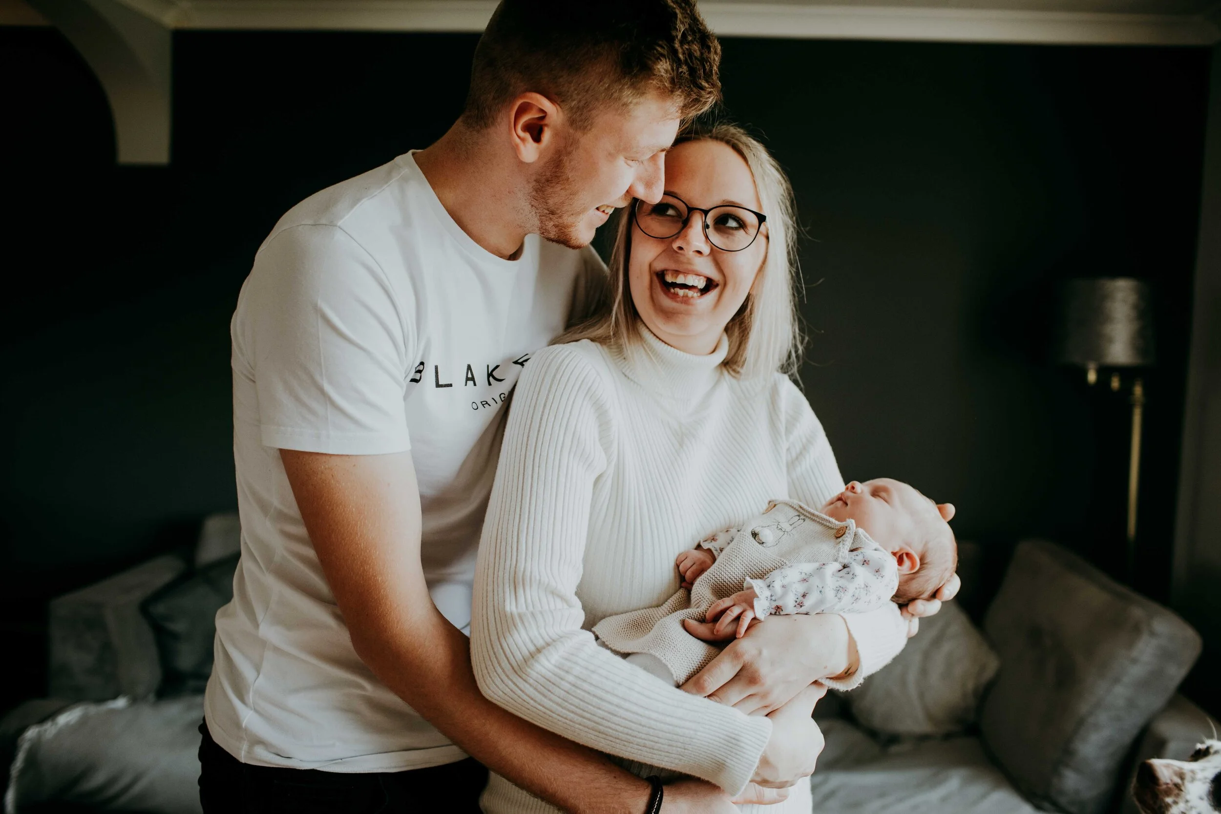 A smiling couple holding a newborn baby in their arms at home, standing against a dark wall with a lamp in the background.