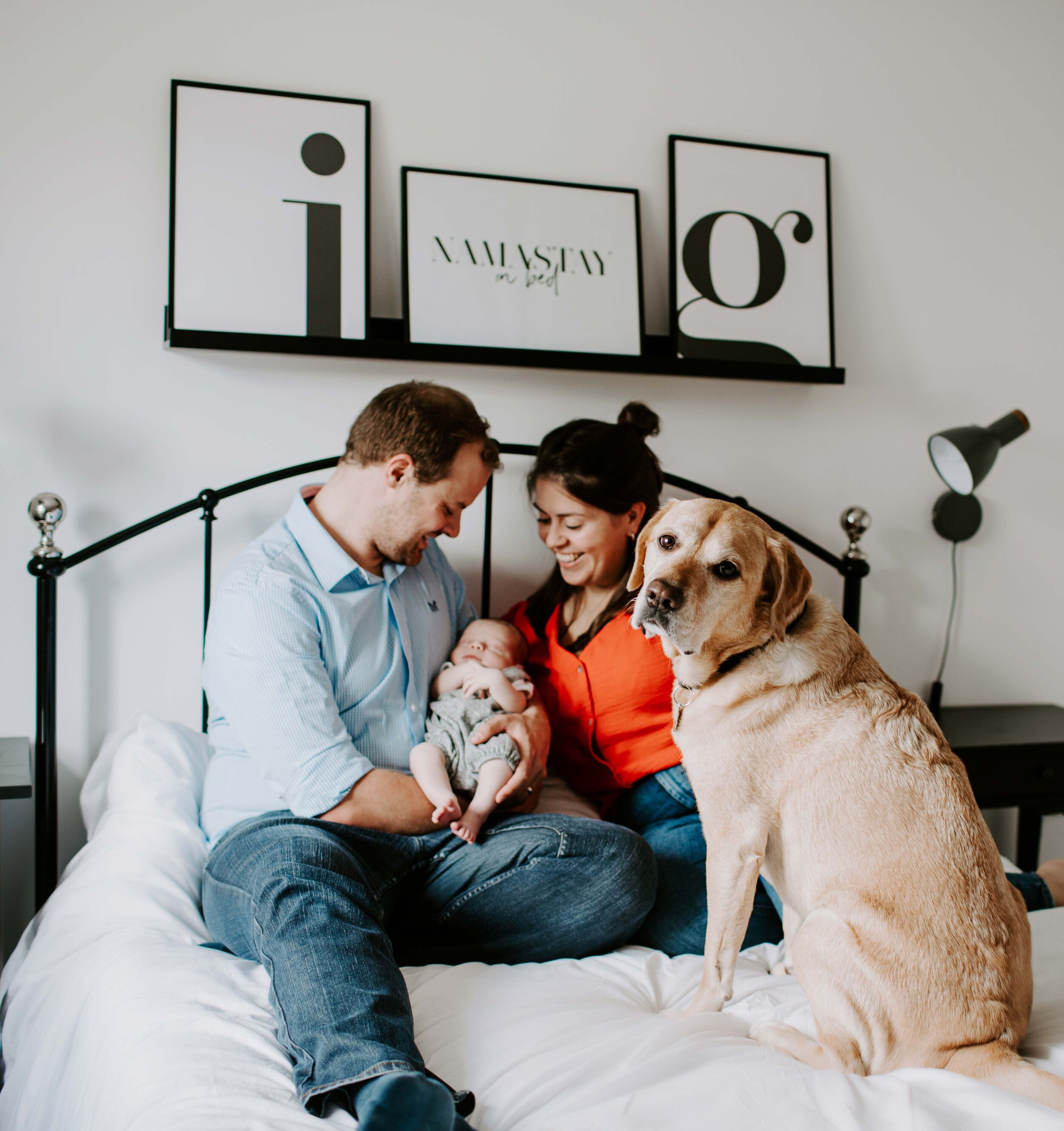 A couple with a baby and a dog sitting on a bed, with framed artwork on the wall in the background.