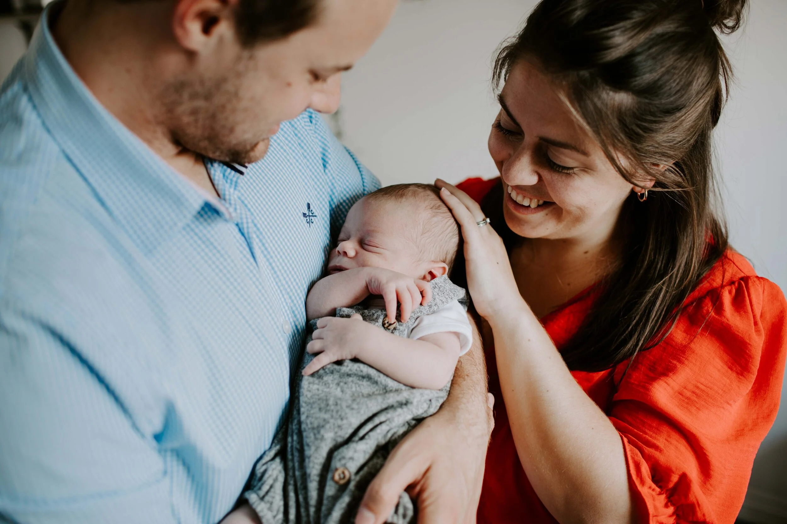 A couple holding a sleeping baby, the man wearing a blue shirt and the woman wearing a red shirt, both smiling and looking at the baby.