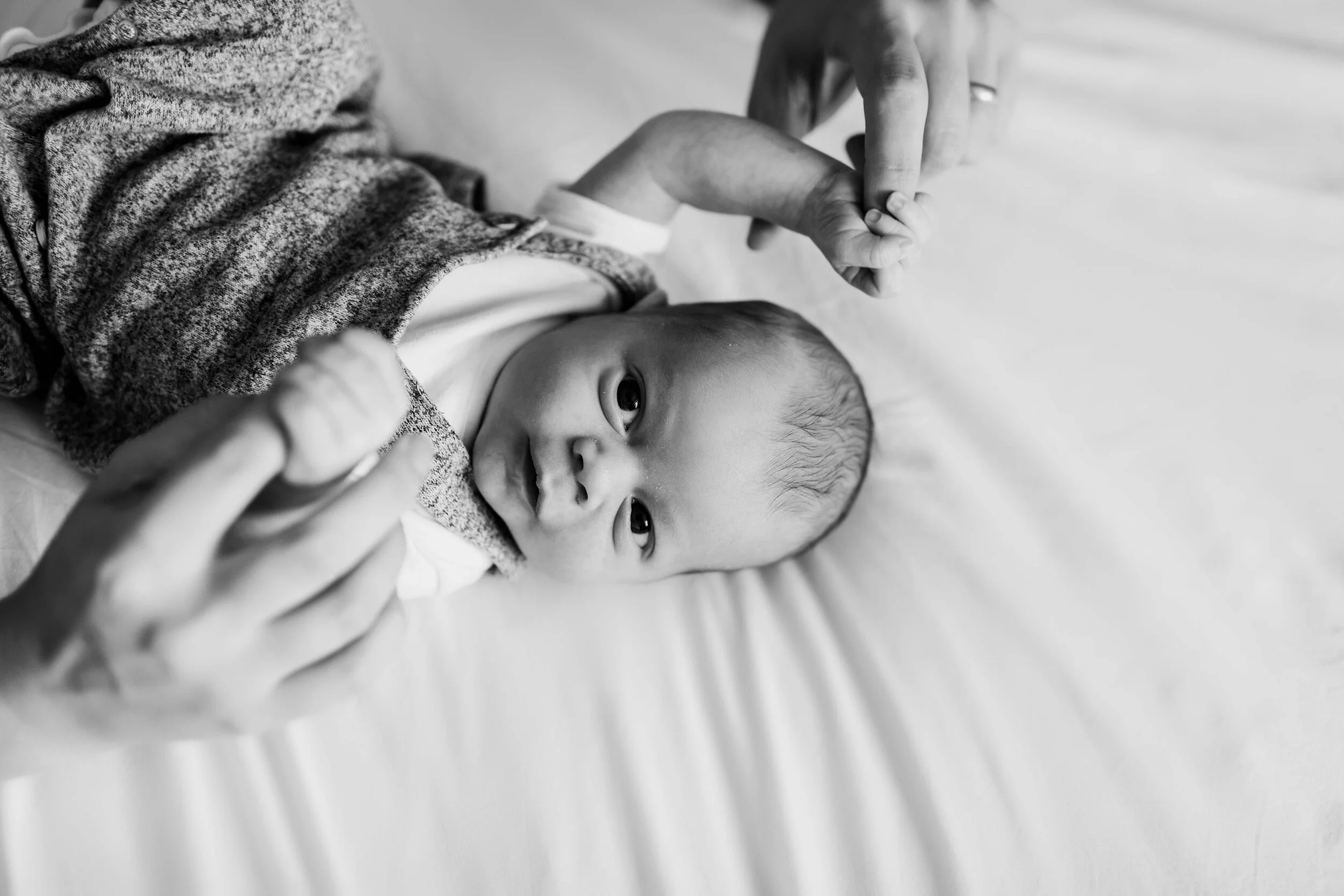 Black and white photo of a baby lying on a bed, holding an adult's hands.
