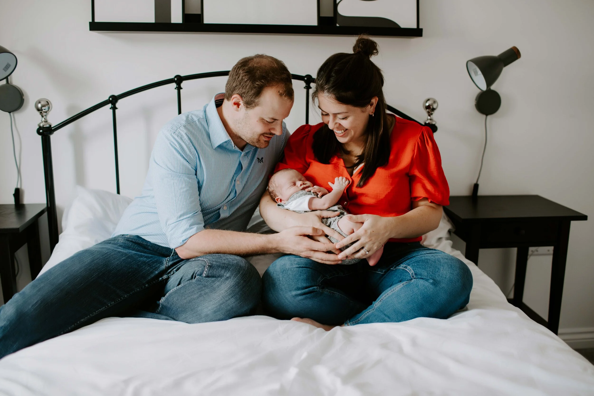 Couple sitting on a bed holding a newborn baby, smiling and looking at the child.
