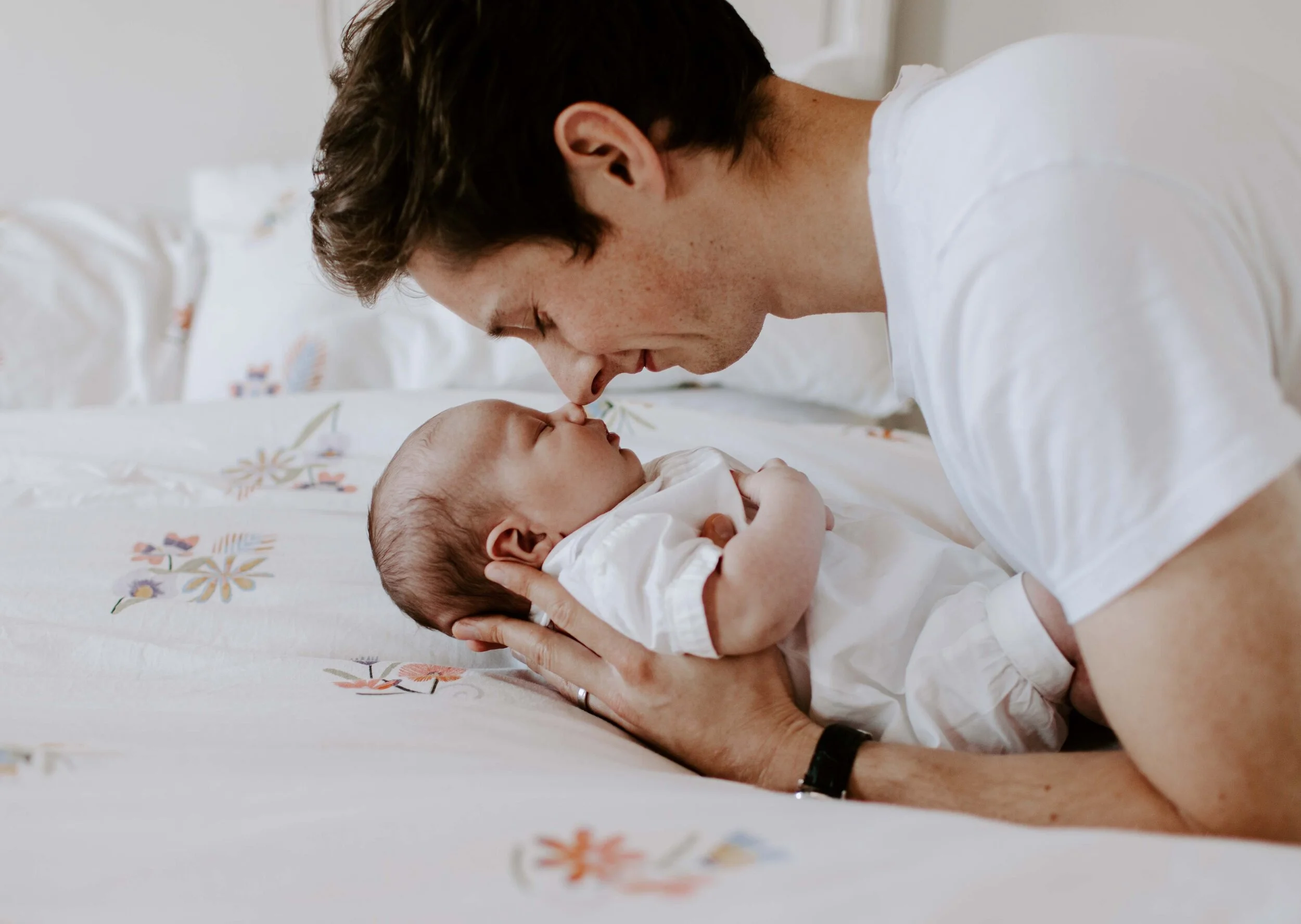 A man gently holds and nuzzles a sleeping newborn baby on a floral bedspread.