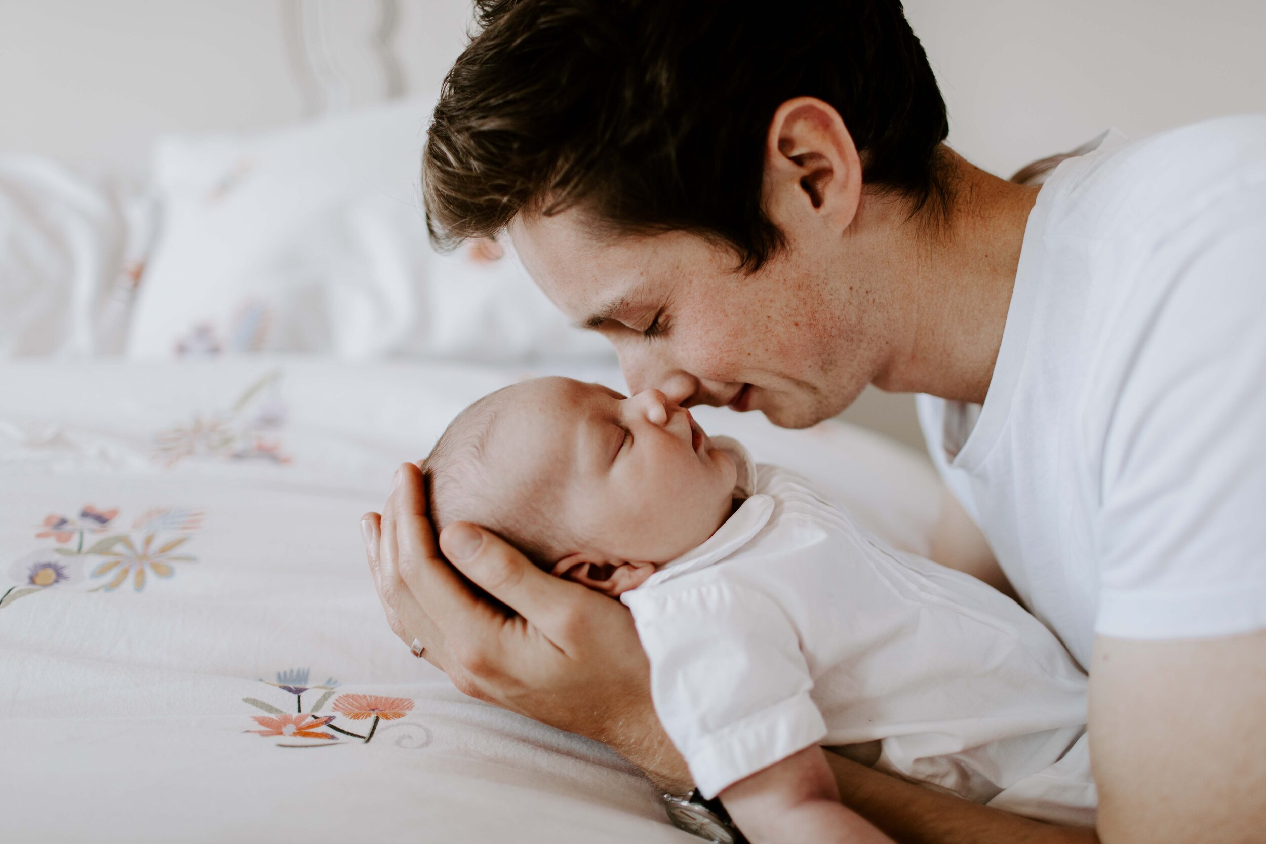 Man gently holding and touching noses with a sleeping baby, both wearing white clothing.