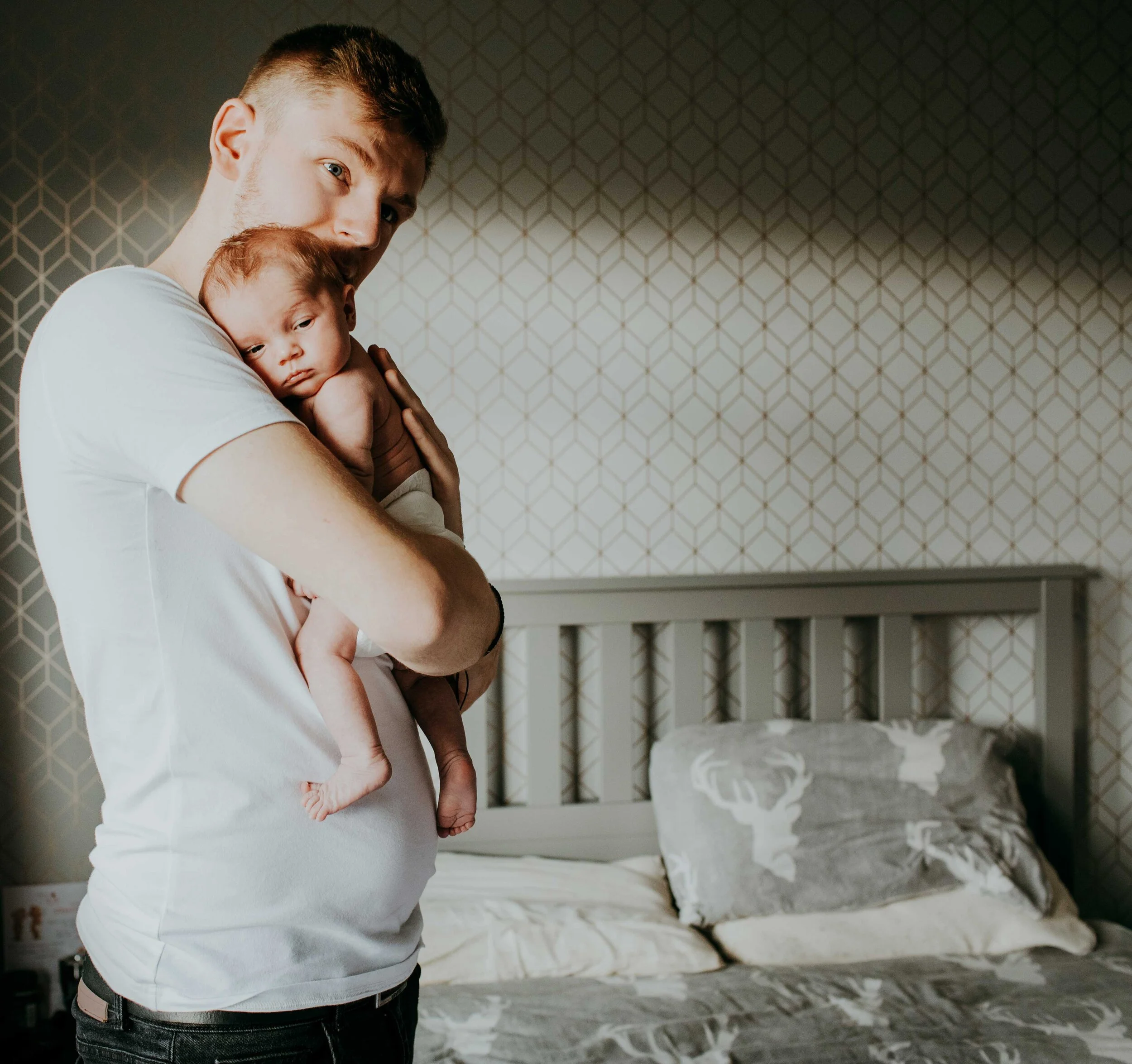 A man holding a baby close to his chest in a bedroom with a patterned wall and bed in the background.