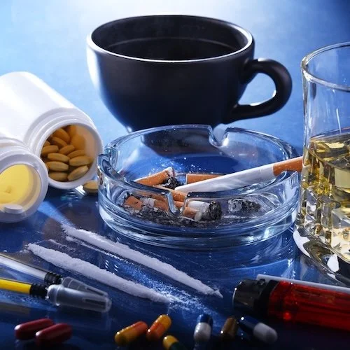 A black coffee mug, an empty pill bottle, and a glass of whiskey surrounded by cigarettes, pills, and a syringe on a blue surface.