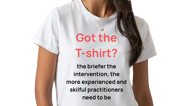 Close-up of a woman wearing a white T-shirt with red and black text about briefer intervention, with long brown hair and a gold necklace.
