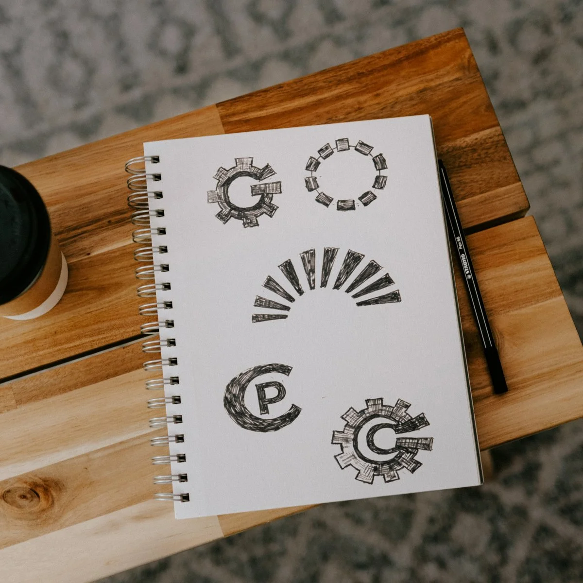 Overhead photo of a sketchbook on a wooden table showing early hand-drawn logo concepts for PlantCore Engineering, including cog shapes and circular marks drawn in black pen, with a pencil and takeaway coffee cup beside it.