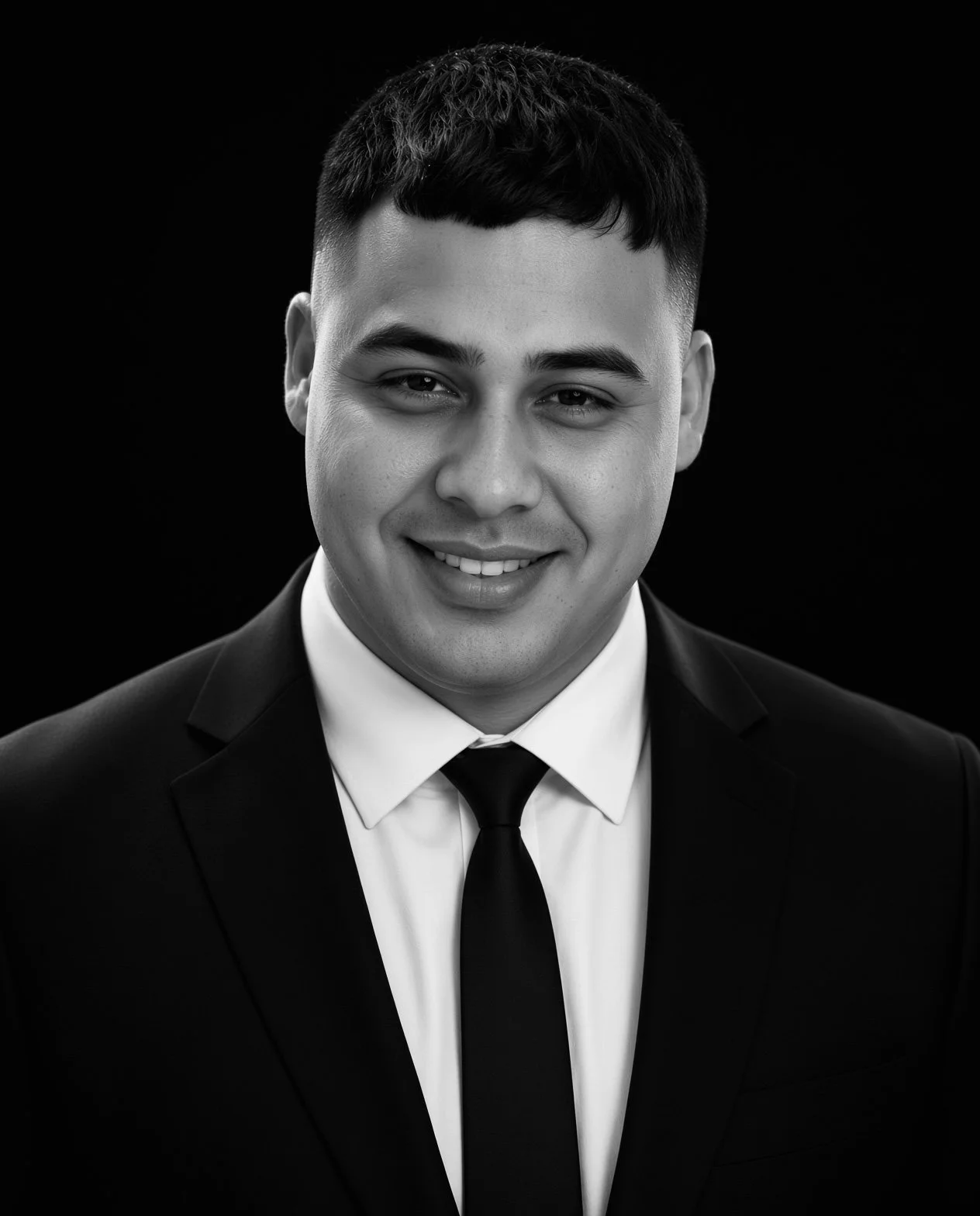 Black and white portrait of a young man wearing a suit, white shirt, and tie, smiling against a dark background.
