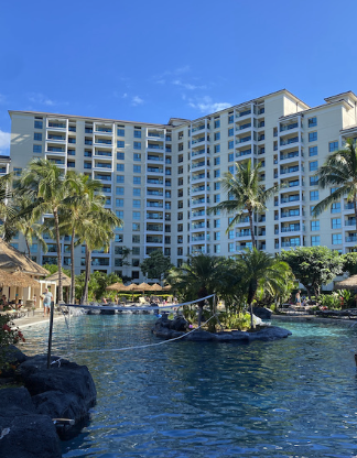 Luxury hotel or resort with a large swimming pool, palm trees, and a high-rise building in the background on a sunny day.