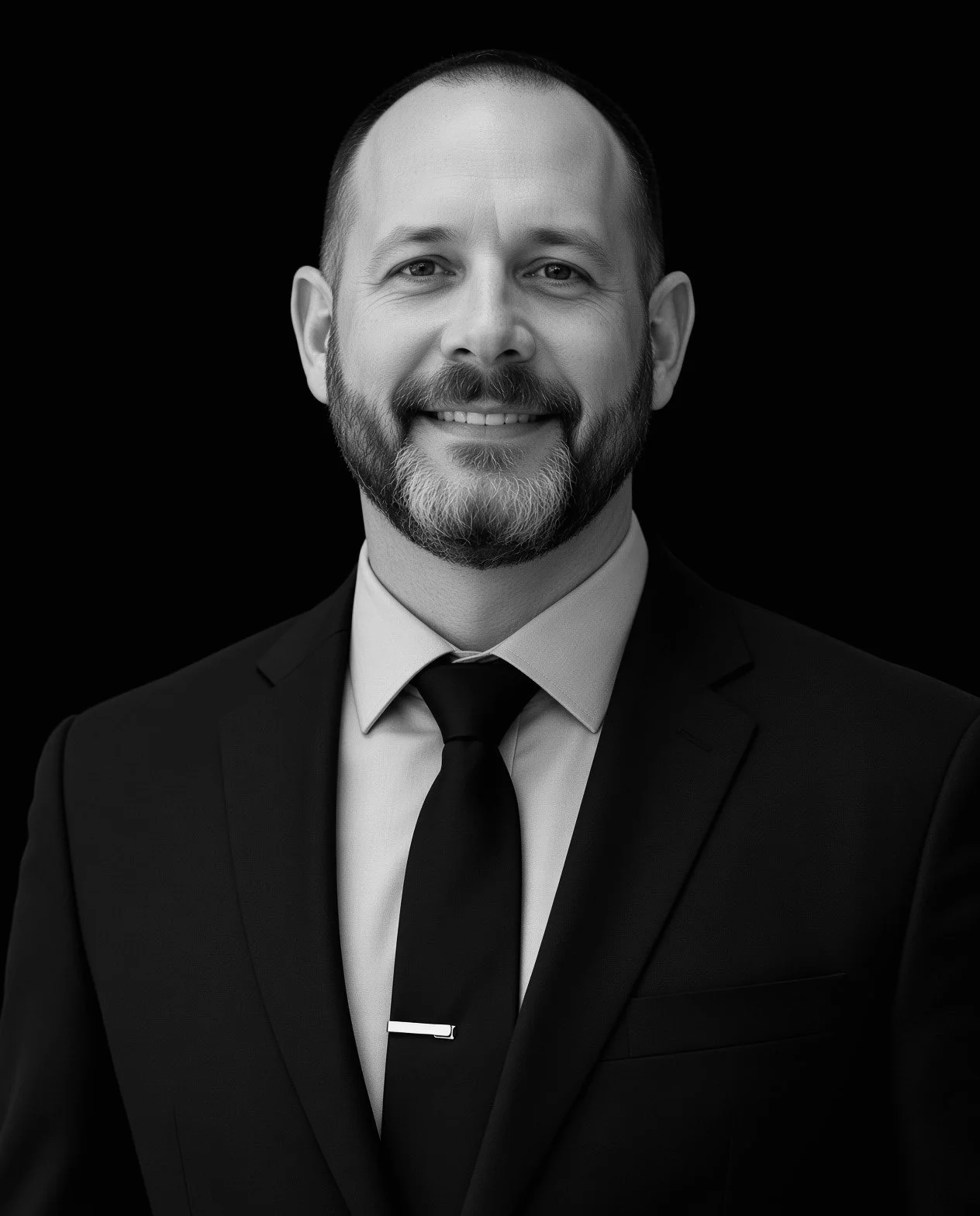 Black and white portrait of a smiling man in a suit and tie, with a beard, against a dark background.