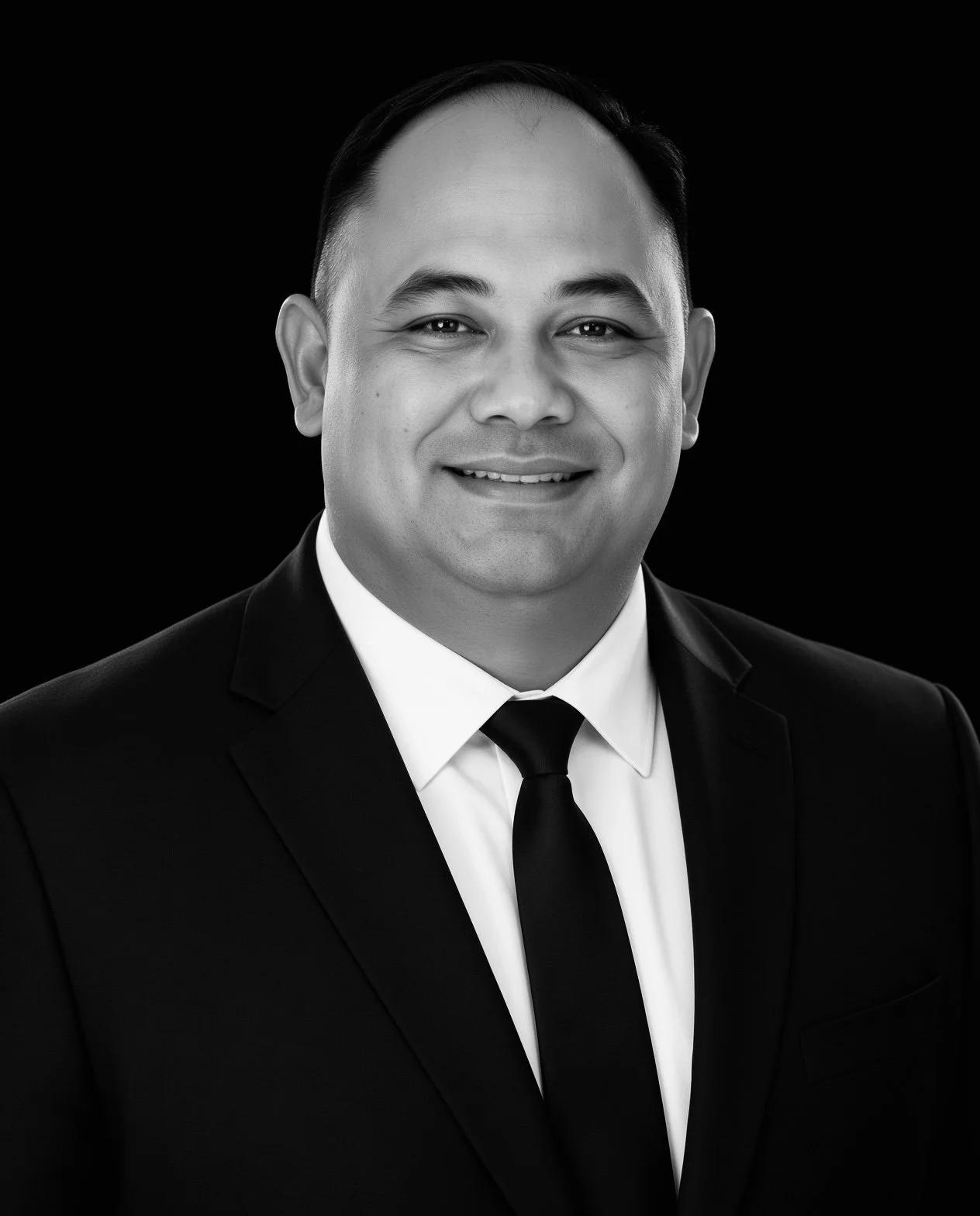 Black and white professional headshot of a smiling man in a suit and tie with a dark background.
