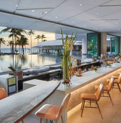 Modern bar with a white marble countertop, tall floral arrangement, and beige seats, overlooking a pool and palm trees at sunset.