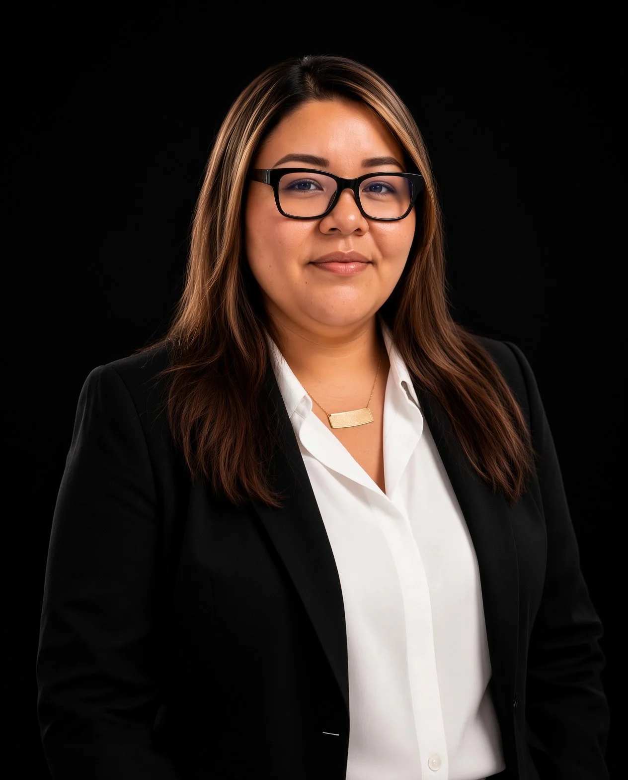 Portrait of a woman with long brown hair wearing glasses, a white shirt, a black blazer, and a gold nameplate necklace against a black background.