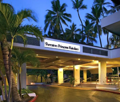 Entrance of the Sheraton Princess Kaiulani hotel surrounded by tall palm trees and lush greenery during early evening.