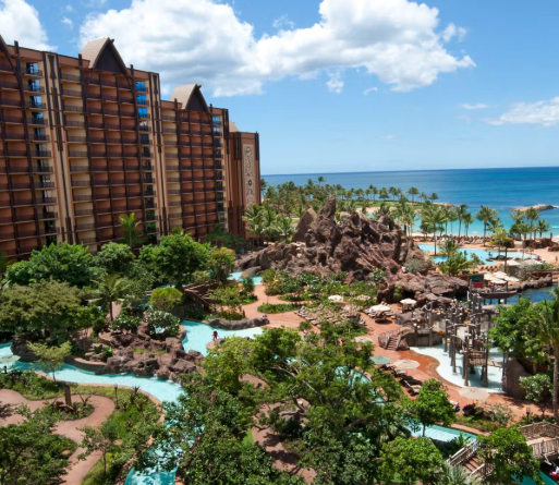 A view of a resort with a large hotel building, lush green landscaping, swimming pools, rocky formations, palm trees, and the ocean in the background.