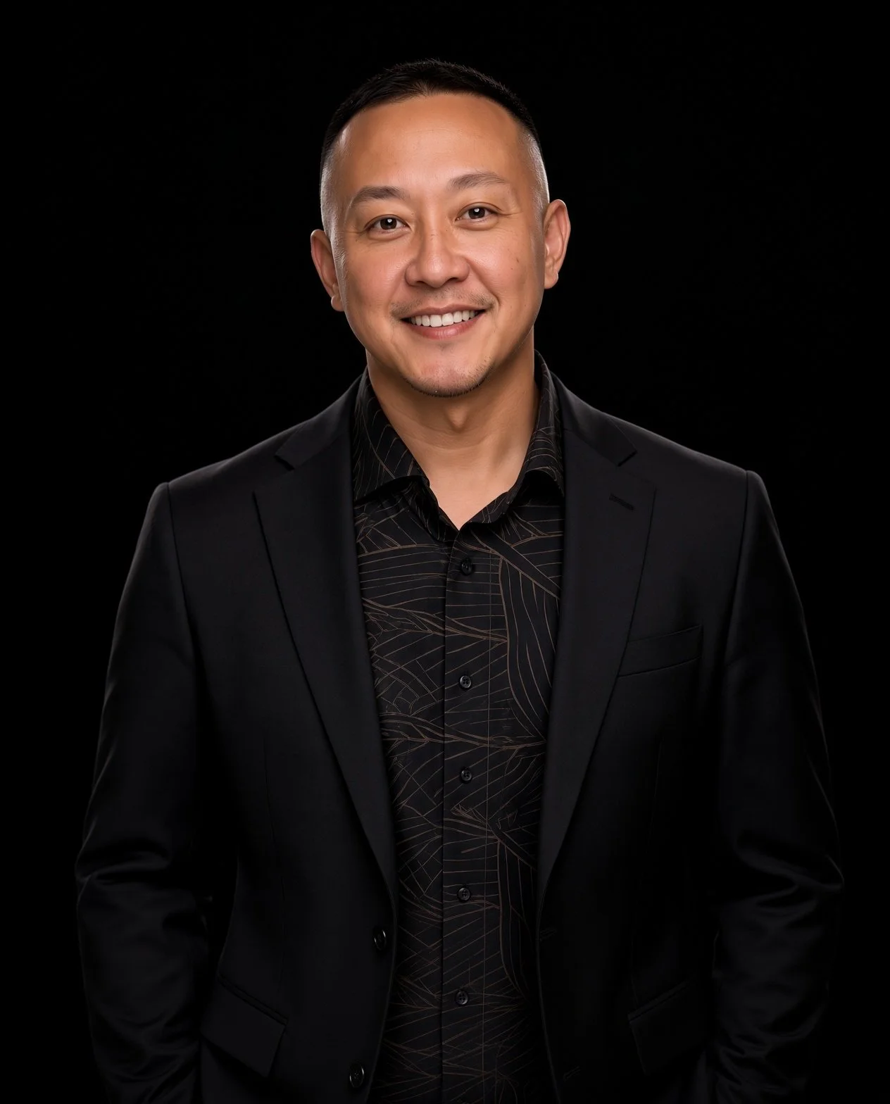 A man with short dark hair, wearing a black blazer over a patterned black shirt, smiling against a black background.