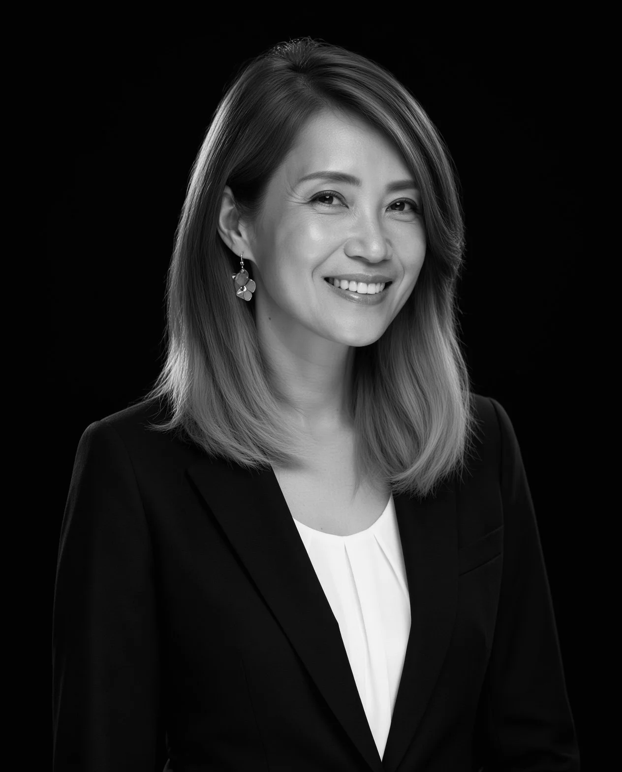 Black and white professional portrait of a smiling Asian woman with shoulder-length hair, wearing a blazer, white blouse, and earrings, against a dark background.