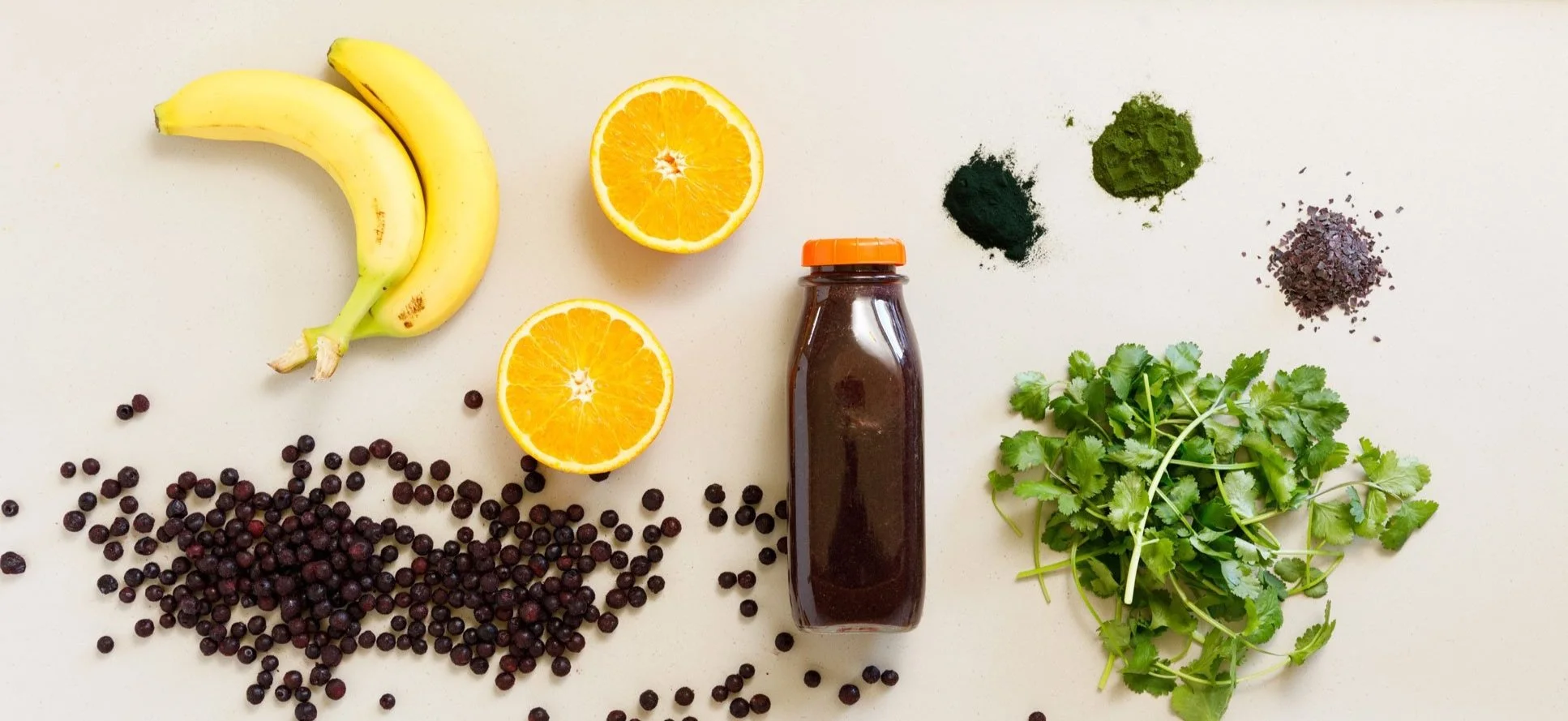 Flasche mit Blaubeersmoothie auf Arbeitsplatte und Zutaten daneben: Bananen, Blaubeeren, Organgen, Koriander, zwei grüne Pulver