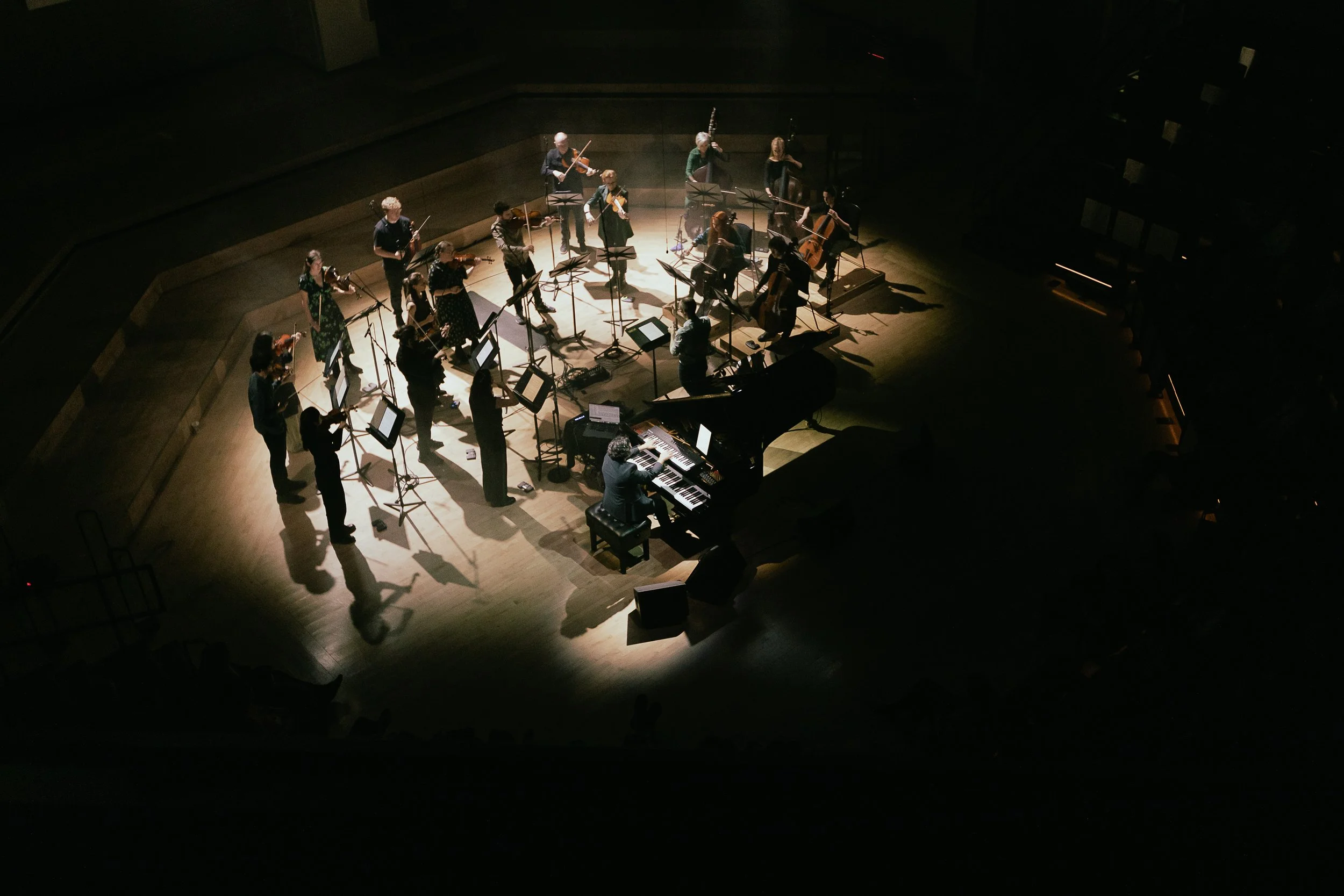 The camera looks down on a performing ensemble from a high angle, soloist seated at a black piano, and a group of standing string instrument players. The lighting is dramatic, with clear pools of light on the performers.