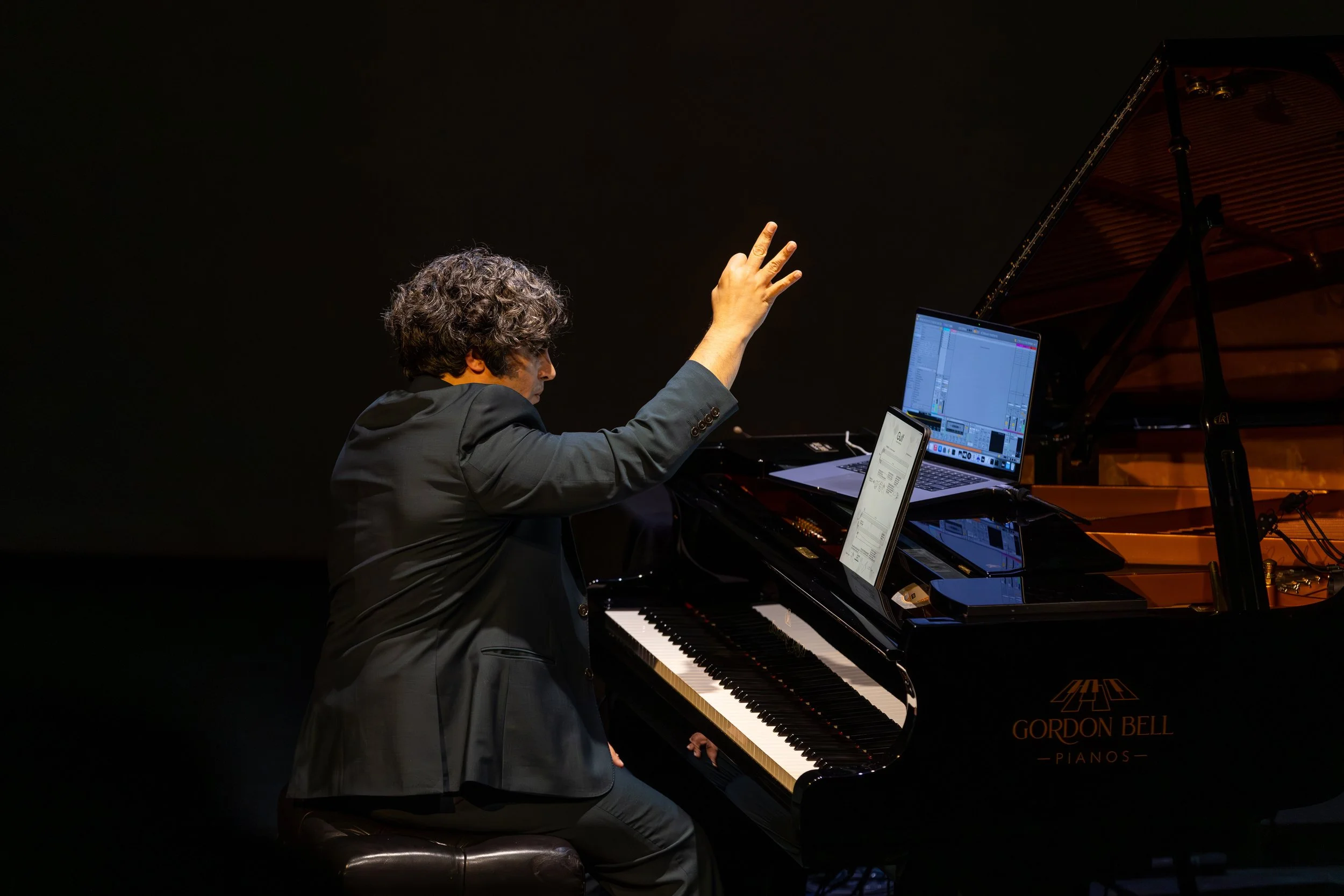 A man with black curly hair wearing a black suit sits at a piano, holding his right hand up, showing a gesture to the webcam of a laptop perched on top of the piano