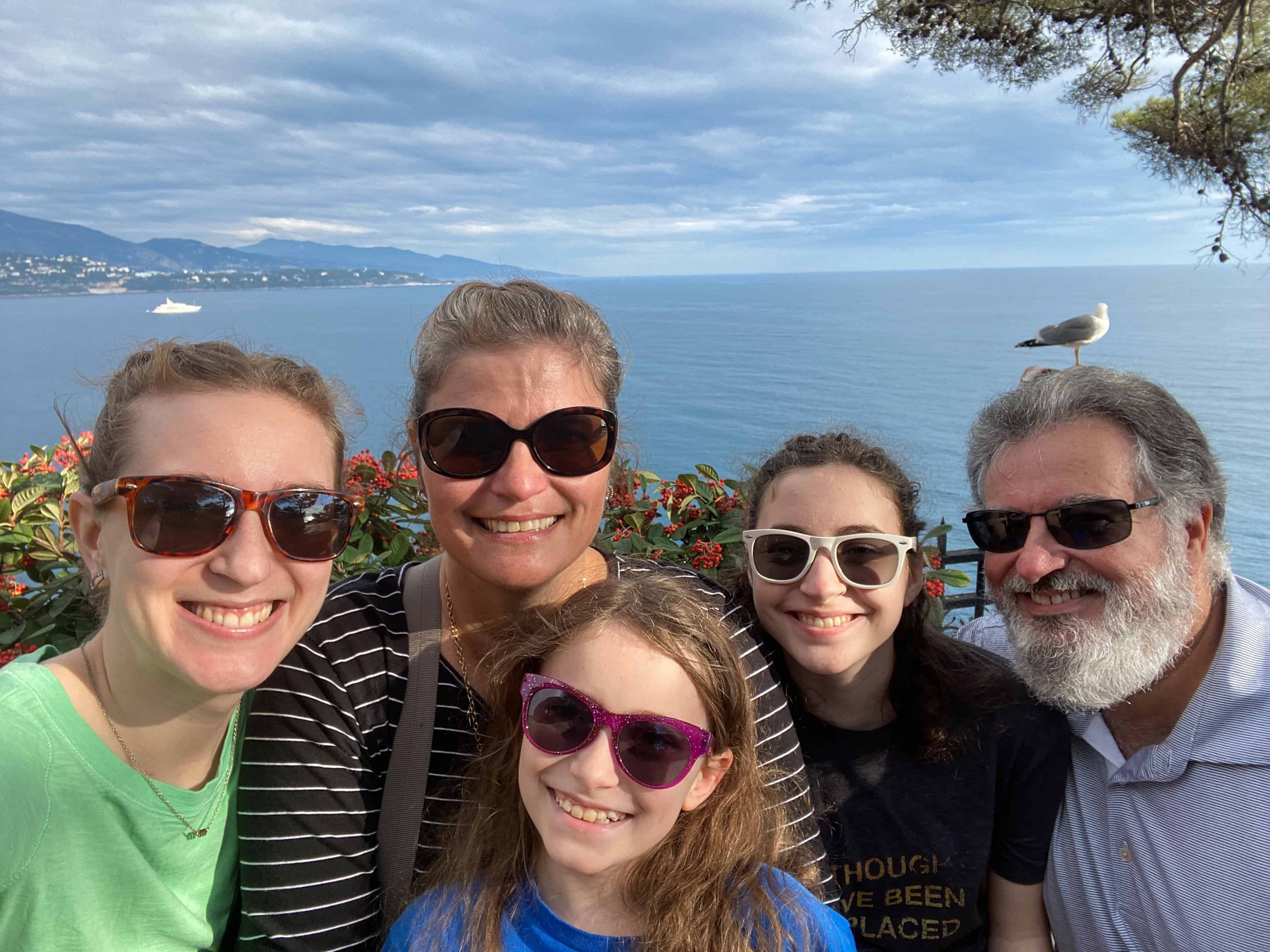 Multigenerational family of five smiling in a selfie above the Mediterranean Sea in Monaco, with a cruise ship, blue water, and a seagull photobombing in the background
