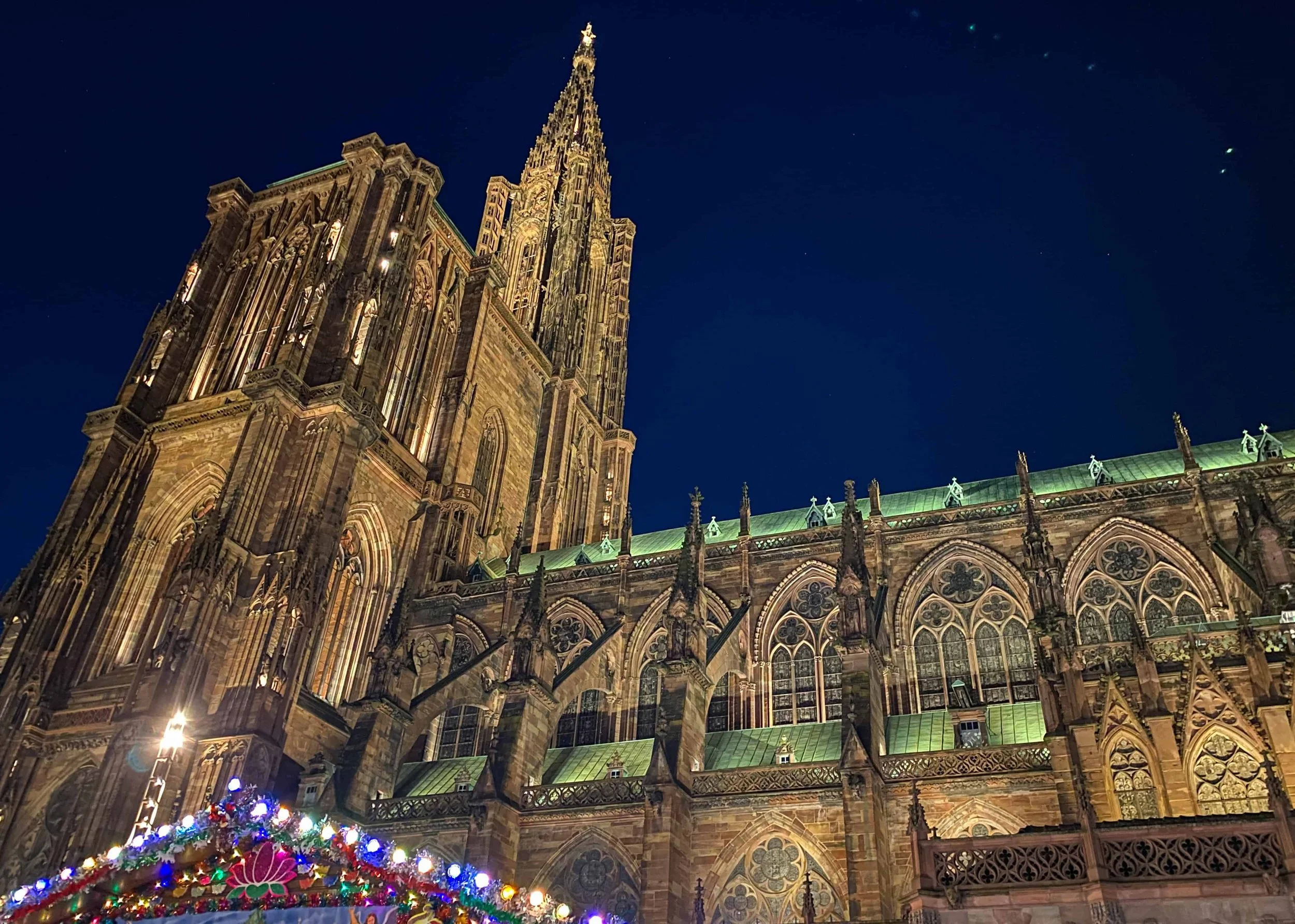 Side view of Strasbourg Cathedral at night with a lit Christmas market chalet in the foreground