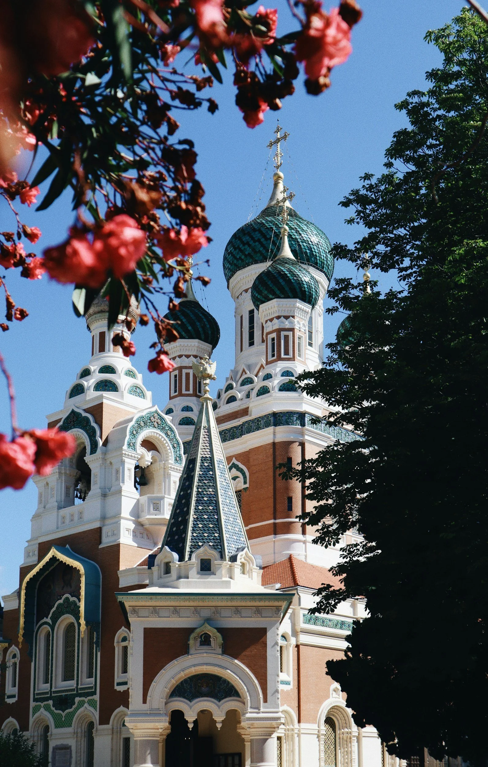 Russian Orthodox Cathedral of Saint Nicholas in Nice, France — green onion domes and ornate brick facade framed by red flowering trees.