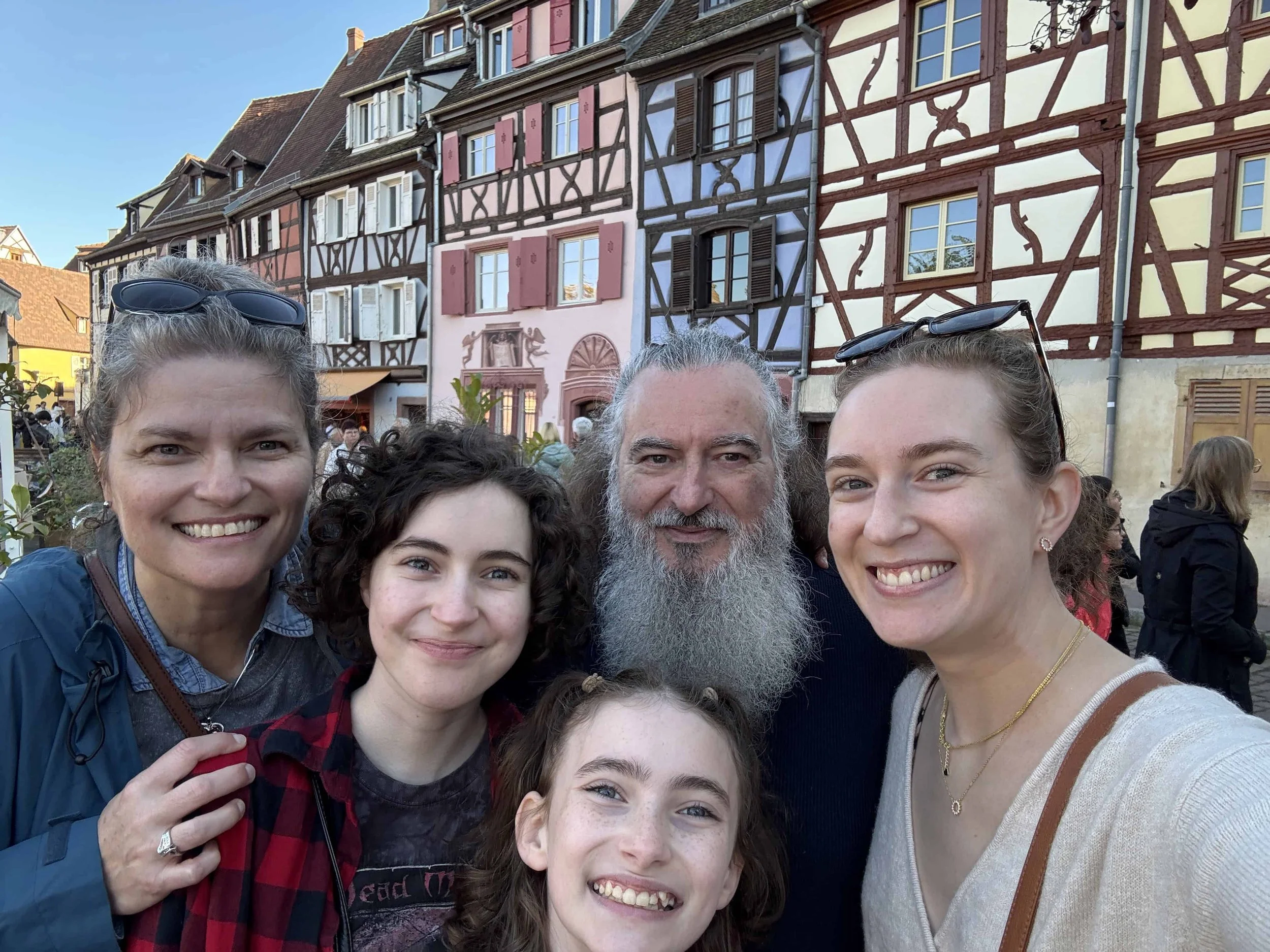 Family of five smiling and taking a selfie in front of colorful, traditional European half-timbered houses in Colmar, France.