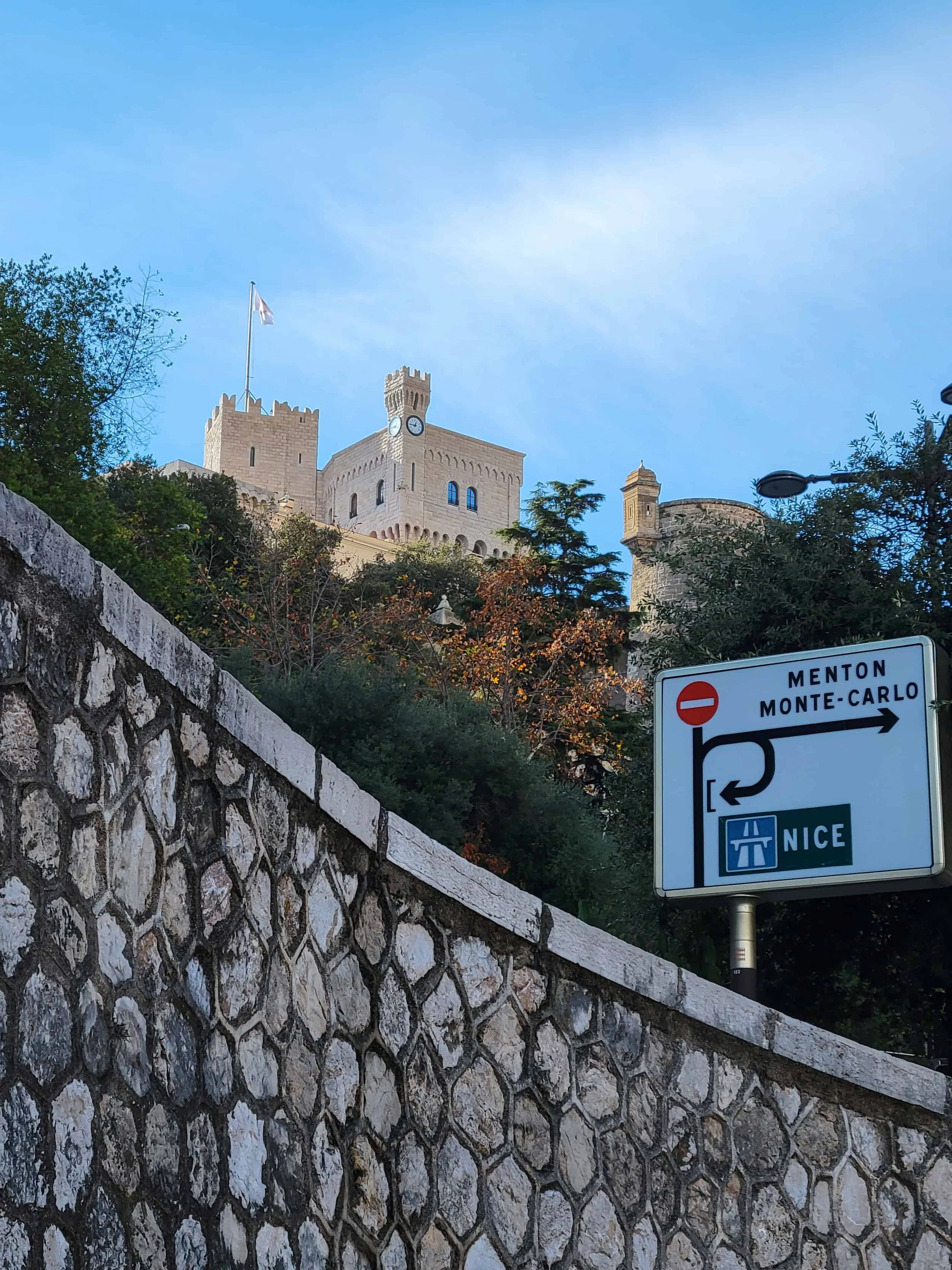 Alt text:  Road sign pointing to Menton, Monte-Carlo, and Nice in front of a stone wall with the Prince's Palace of Monaco rising on the rock above — the view from near Port Hercule on a Monaco day trip from Nice