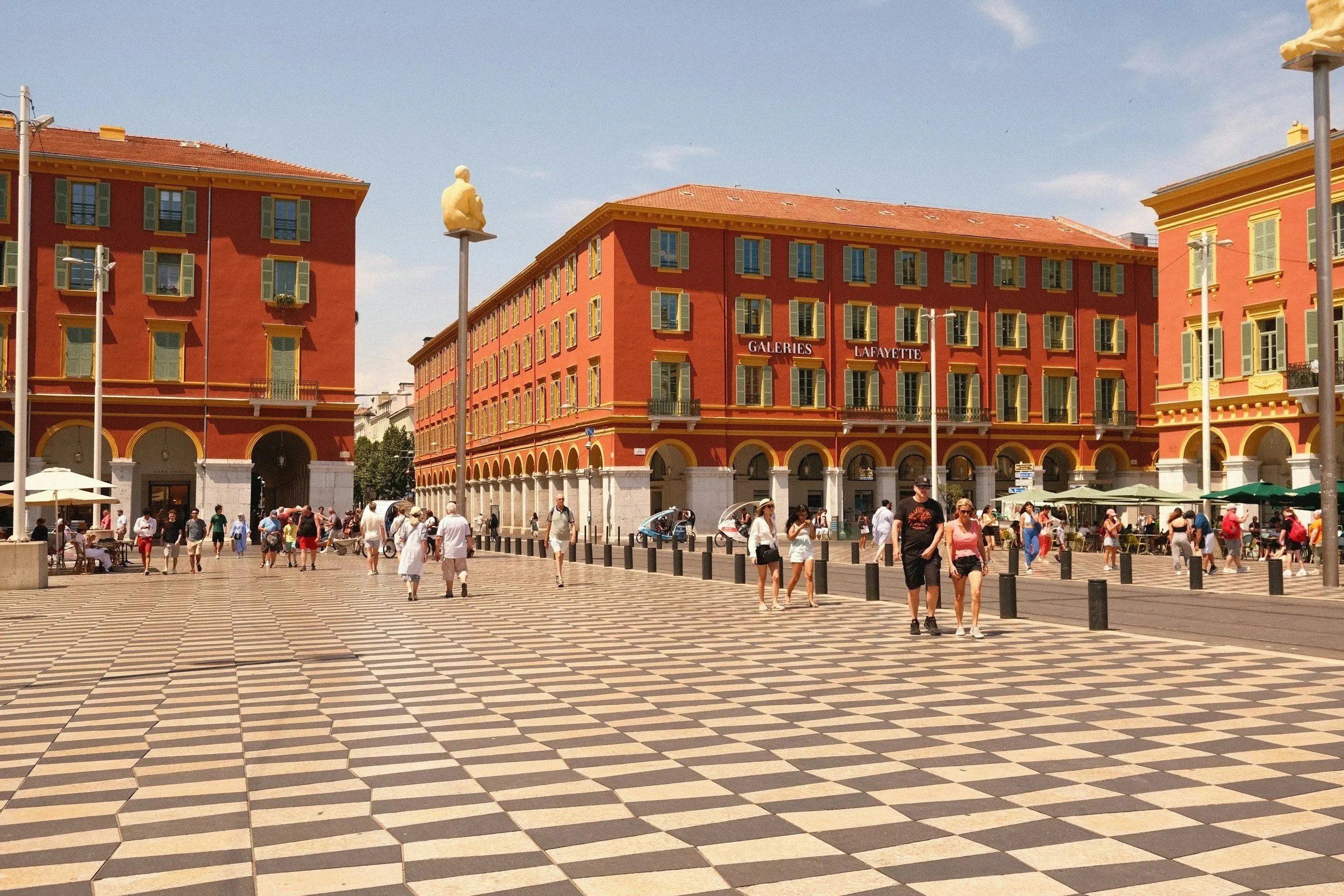 Place Masséna in Nice, France — black and white checkerboard pavement, ochre-red buildings, Galeries Lafayette, and one of Jaume Plensa's illuminated sculptures on a tall pole.