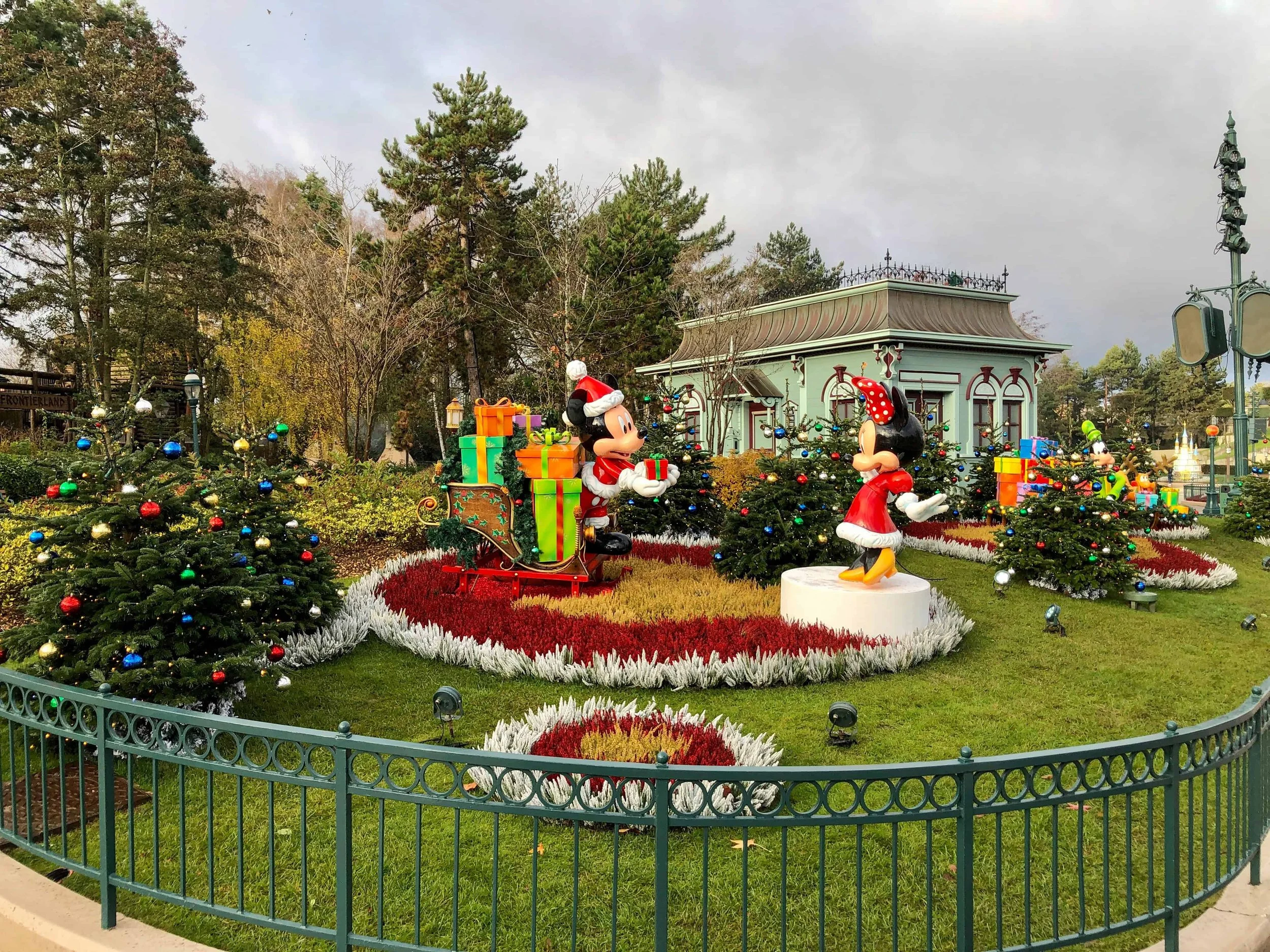 Christmas display of Mickey in a Santa suit handing Minnie a gift, surrounded by decorated trees and red and white flowers.