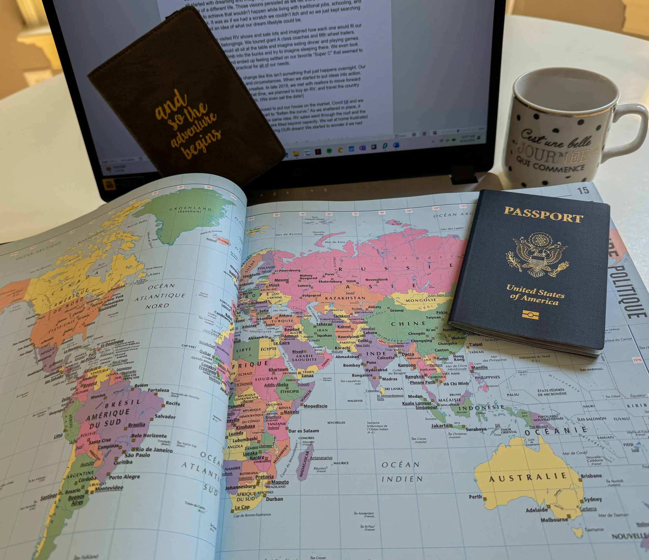 A world map open to Africa, Europe, and Asia. There is a United States passport on the map, a coffee mug, a closed brown notebook with the words 'and so the adventure begins' written on it, and a laptop in the background.