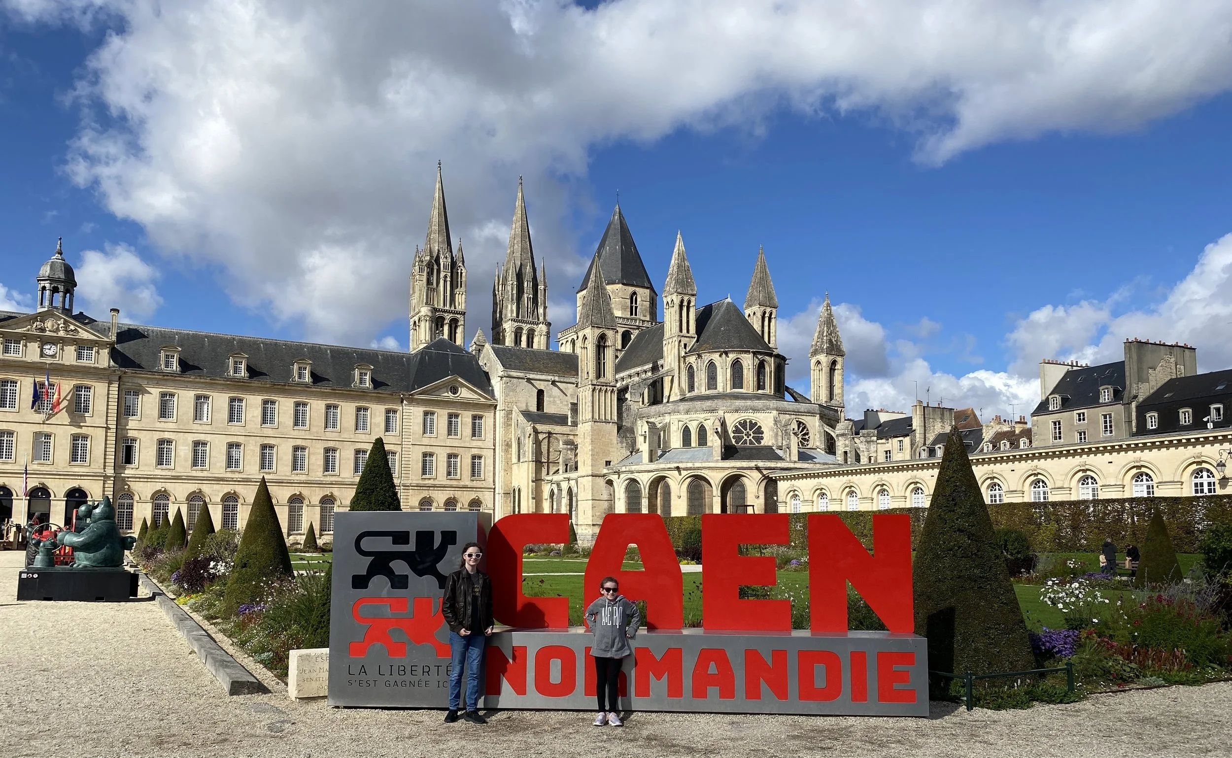 Two family teens posing in front of the Caen, France sign in front of the Abbaye-aux-Hommes, a history lesson for family trips to Normandy in 2026