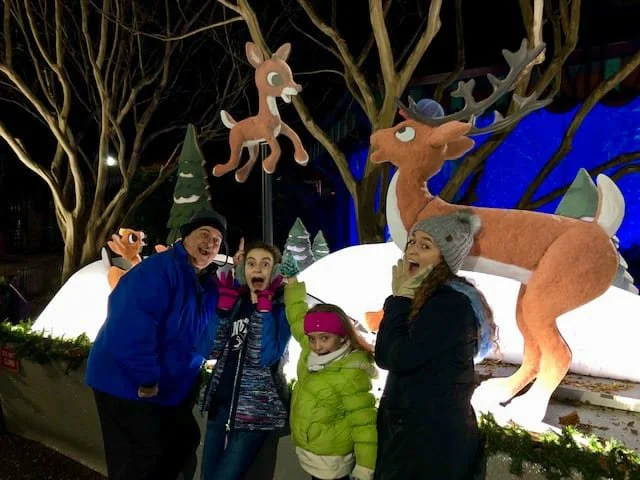 Four family members in winter coats pose in surprise beside reindeer figures, including a flying Rudolph, at night.