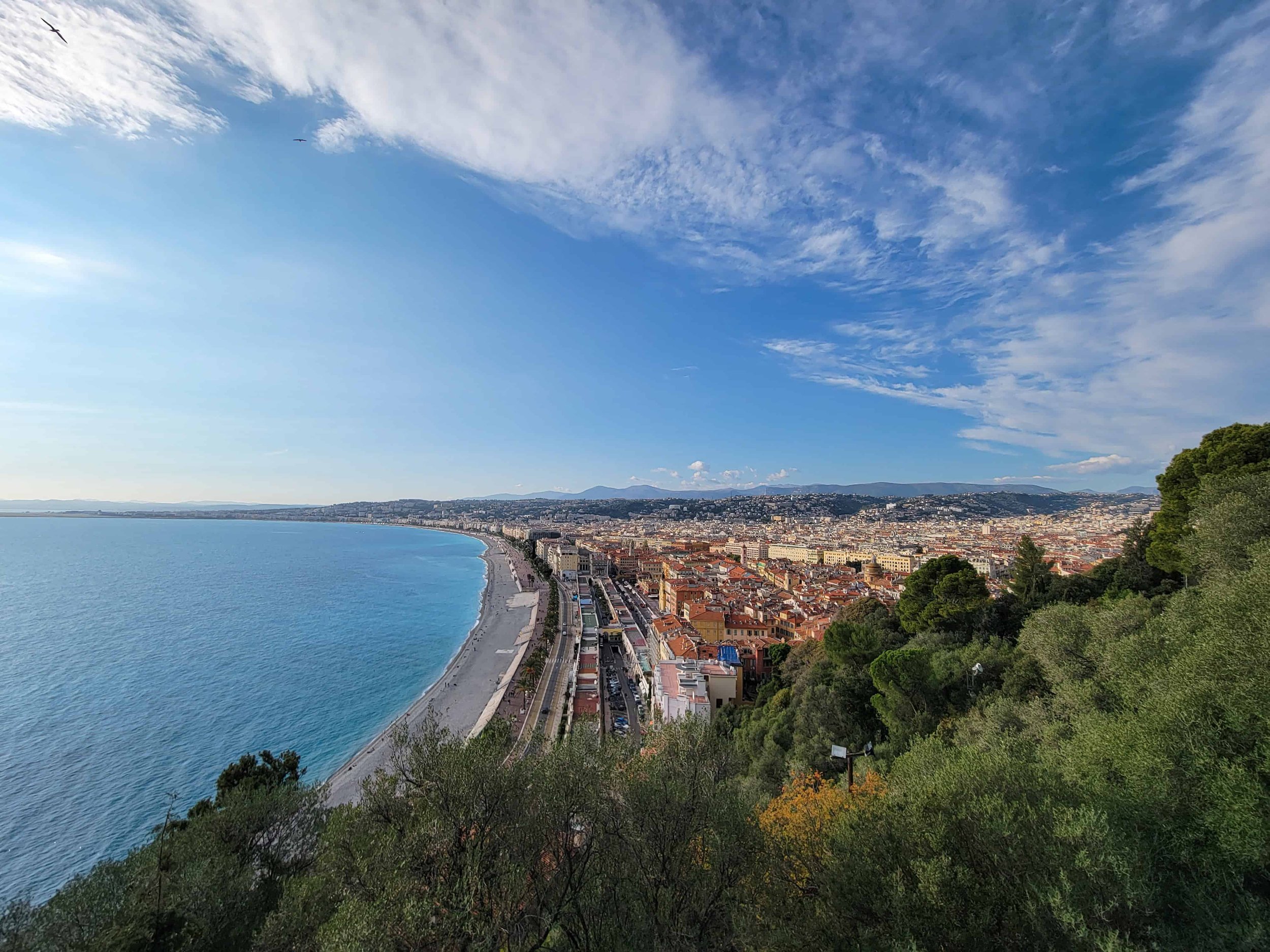 Panoramic view of the Promenade des Anglais and Nice, France from Castle Hill — pebbled beaches, Mediterranean Sea, and the city's terracotta rooftops stretching to the Alps.