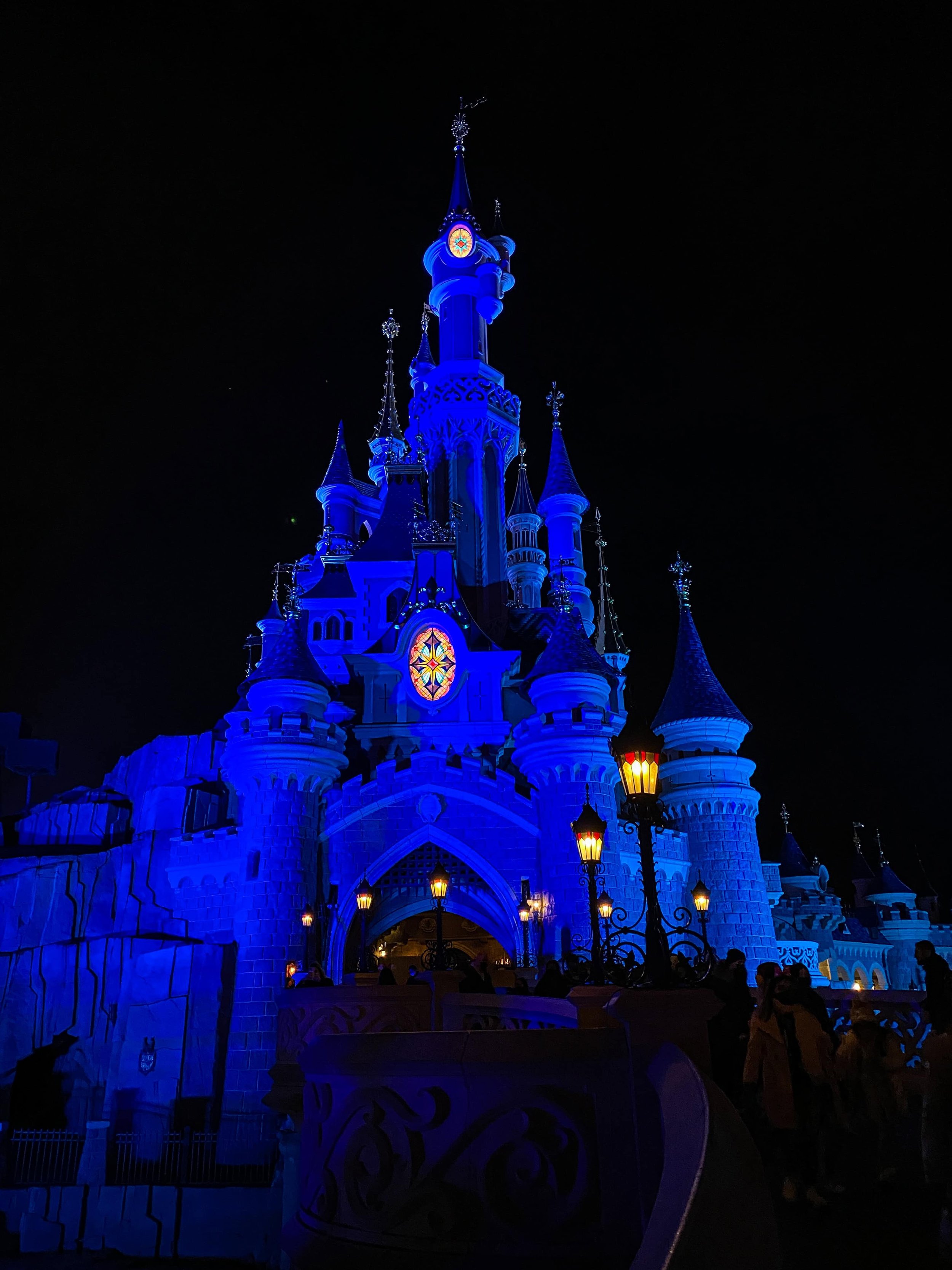 Sleeping Beauty Castle at Disneyland Paris lit deep blue at night, with lanterns along the walkway and guests in silhouette.