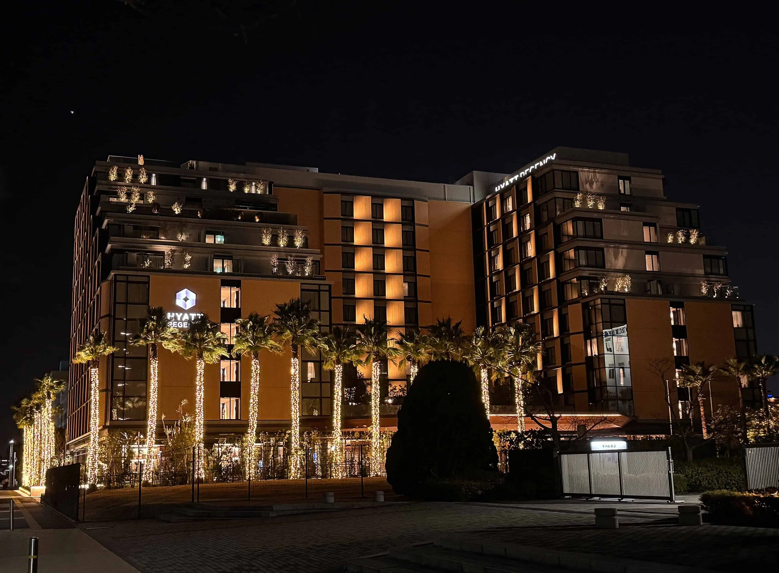 Hyatt Regency Tokyo Bay at night, a tiered hotel with glowing windows and palm trees wrapped in white lights