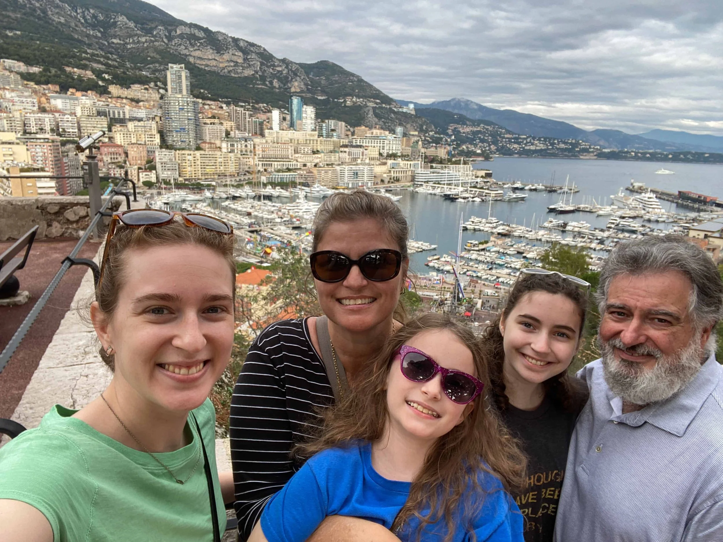 Multigenerational family of five smiling in a selfie above Port Hercule marina in Monaco, with hundreds of superyachts, Monte Carlo's hillside skyline, and the Alps visible behind them