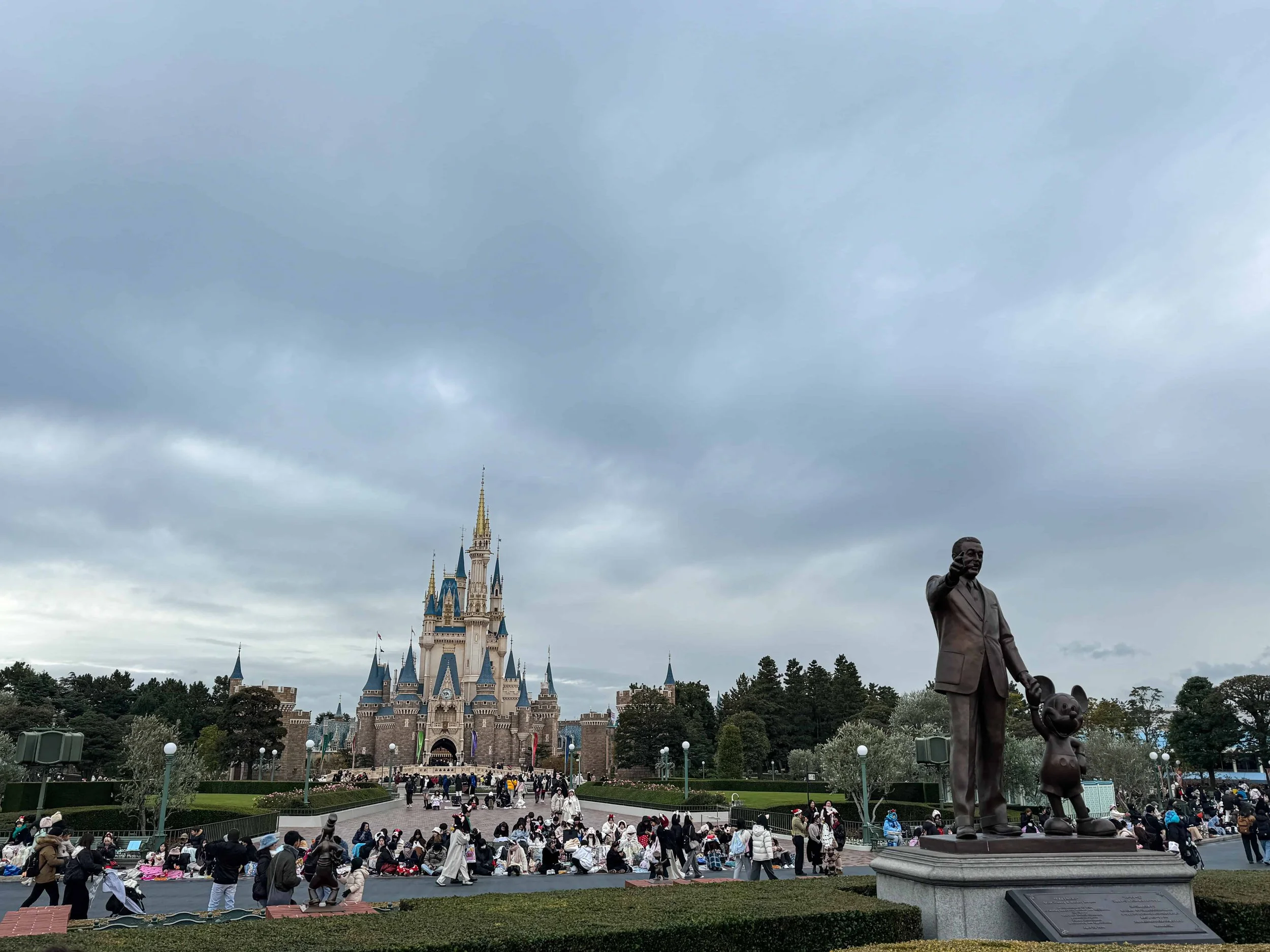 Cinderella Castle in the distance across a plaza of seated guests, with a bronze Walt Disney and Mickey statue at right